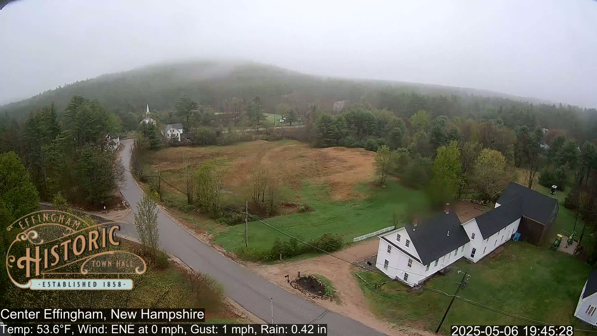 A misty, rainy view of a rural area shows a road leading to a white farmhouse and a church steeple nestled among green trees and brown fields on a hillside.