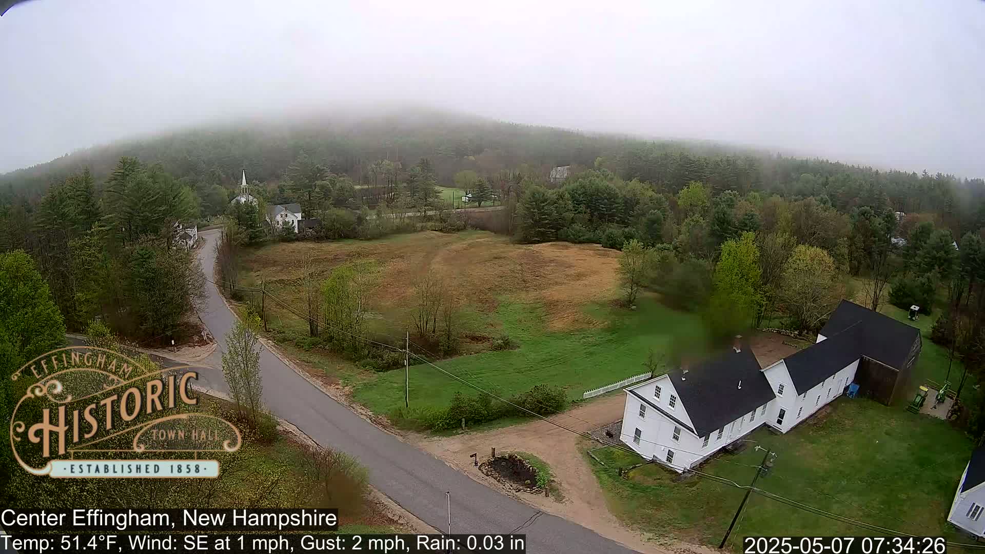 A foggy, lightly rainy scene shows a rural road intersecting with another road near several white buildings and a grassy field, all surrounded by a wooded hillside.