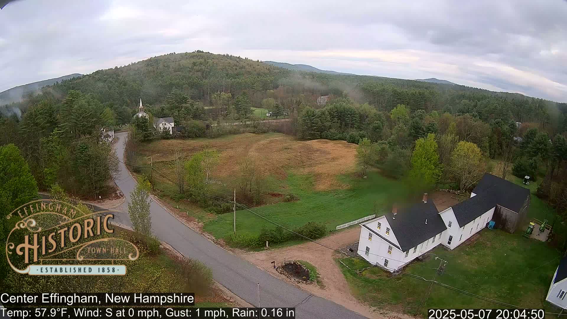An elevated view of a rural area shows a road intersecting near a white house and barn, with a church steeple visible in the distance under an overcast sky, light rain falling.