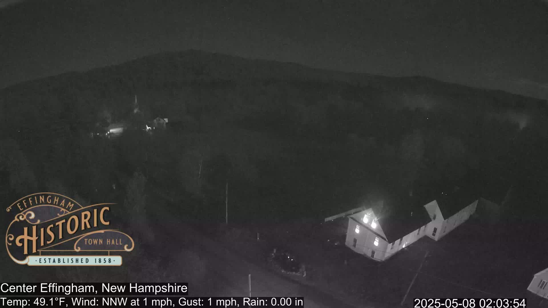 A nighttime, low-light view shows a building and a hill under a clear sky with a slight wind.