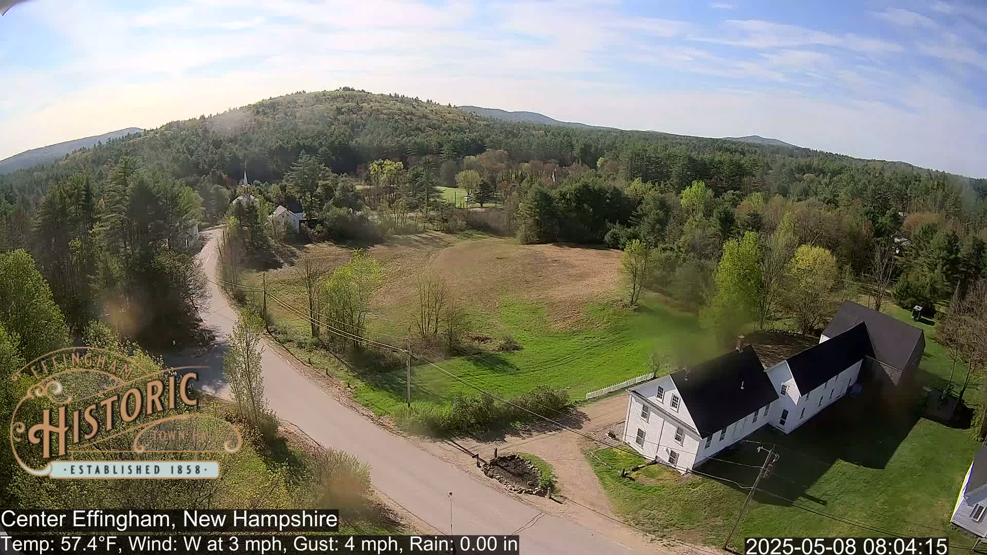An aerial view shows a rural New Hampshire town on a partly sunny day, with a road curving past a white building and toward a forested hill in the distance.