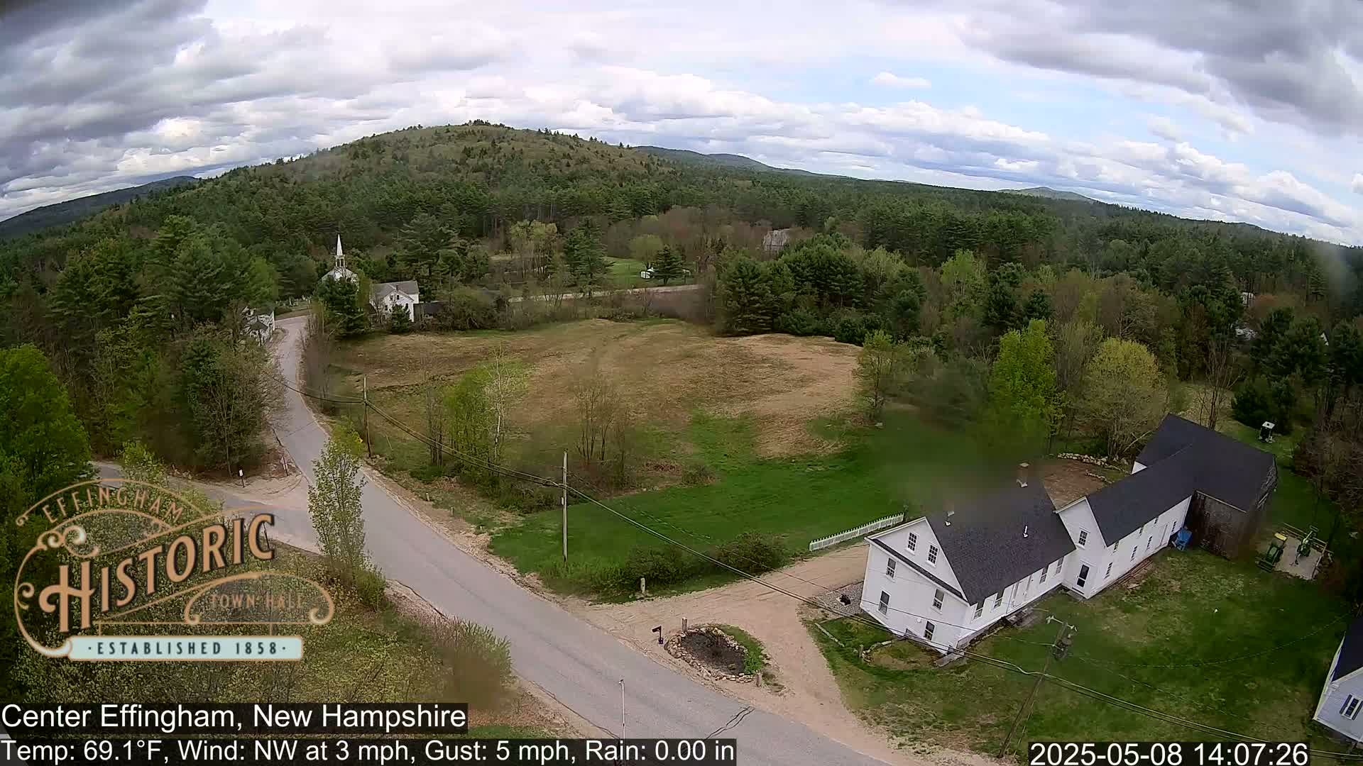 An aerial view shows a rural area with a road, houses, and green trees under partly cloudy skies.