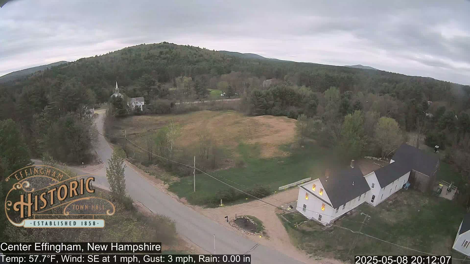 An aerial view shows a small town nestled among green hills under an overcast sky, with a road leading to several buildings, including a white house with lit windows.