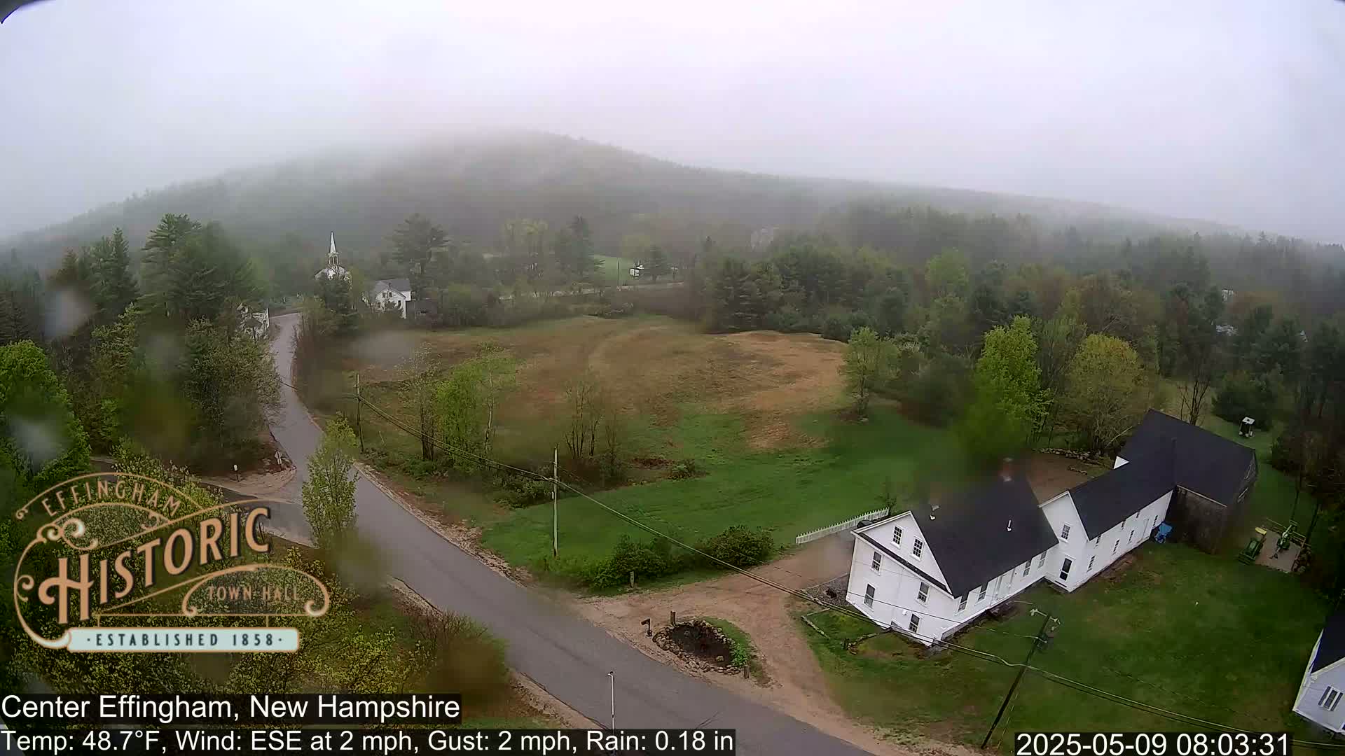 A rain-soaked, elevated view shows a rural area with houses, roads, and green fields, under a misty sky.