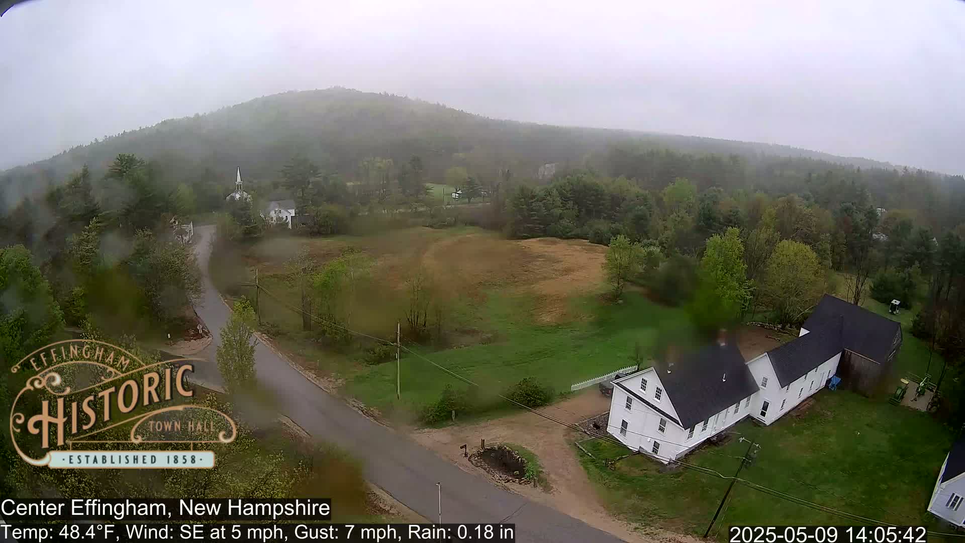 A rain-swept view of a rural area with a white farmhouse, a road, and green hills under an overcast sky.