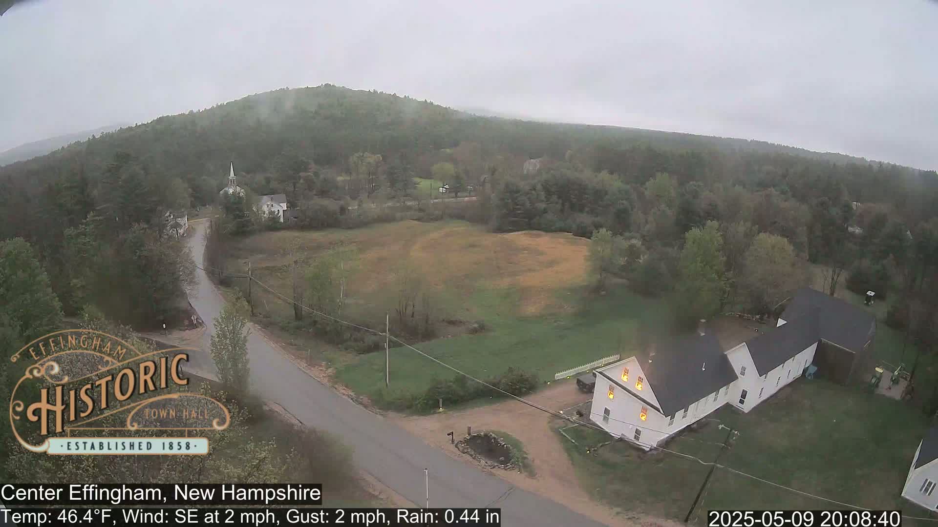 A rainy, overcast view of a small town nestled among green hills, featuring a white farmhouse with lit windows and a church steeple in the distance.