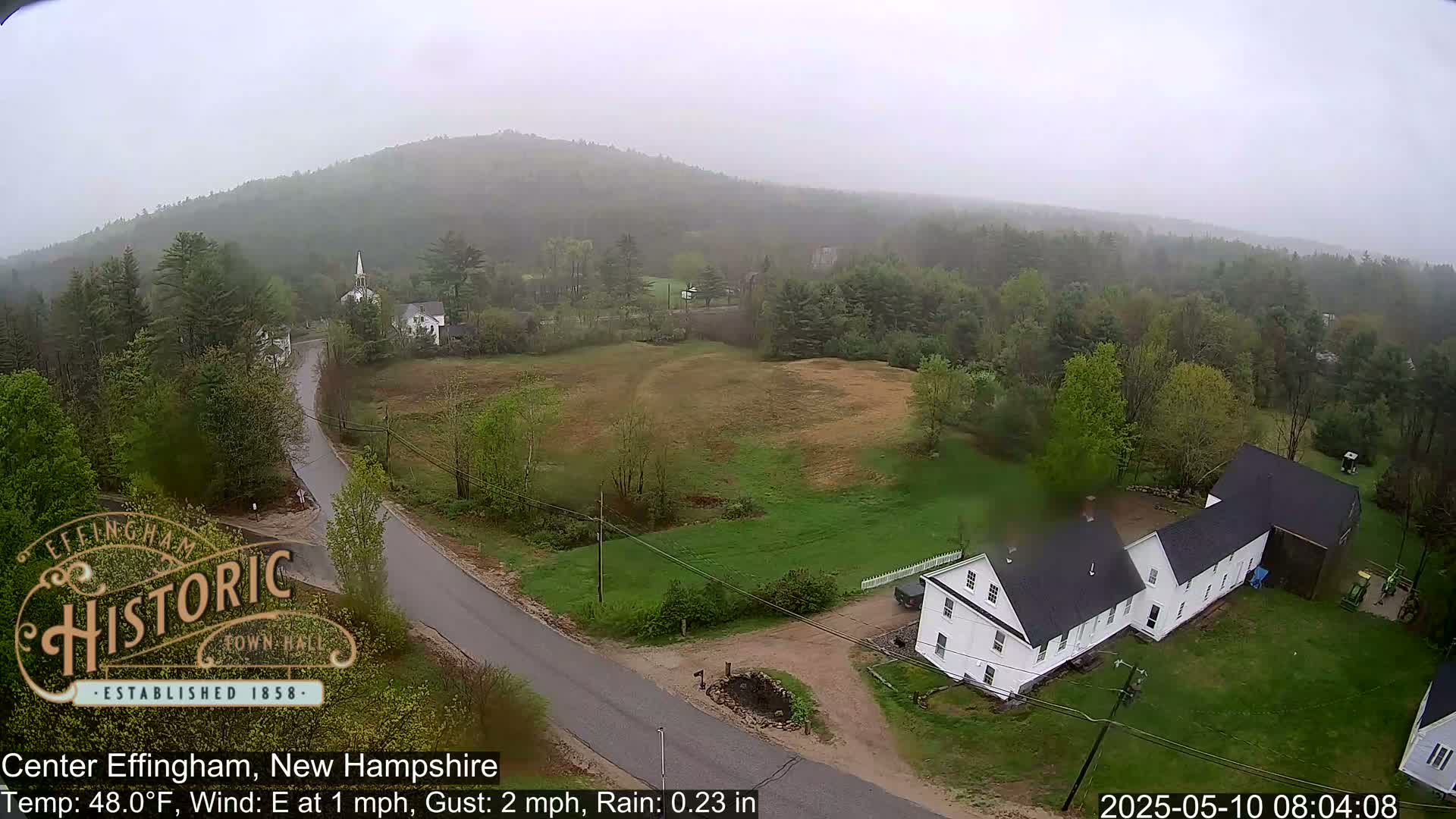 A rain-swept view of a rural New Hampshire town shows a road curving past a white farmhouse and towards a church steeple, all nestled amongst green hills under an overcast sky.