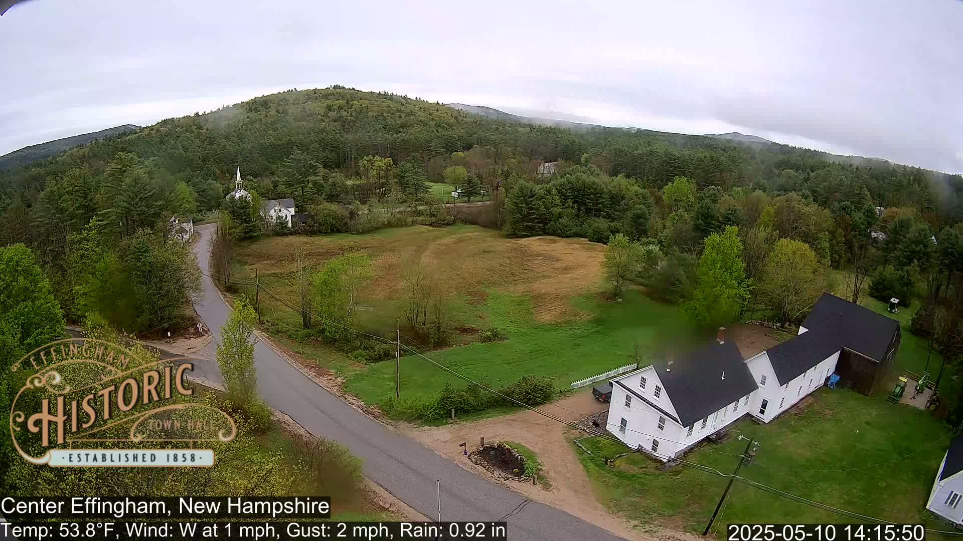 A rainy, overcast day reveals a small New England town nestled amongst green hills and trees, with a white church steeple and several houses visible from an aerial perspective.