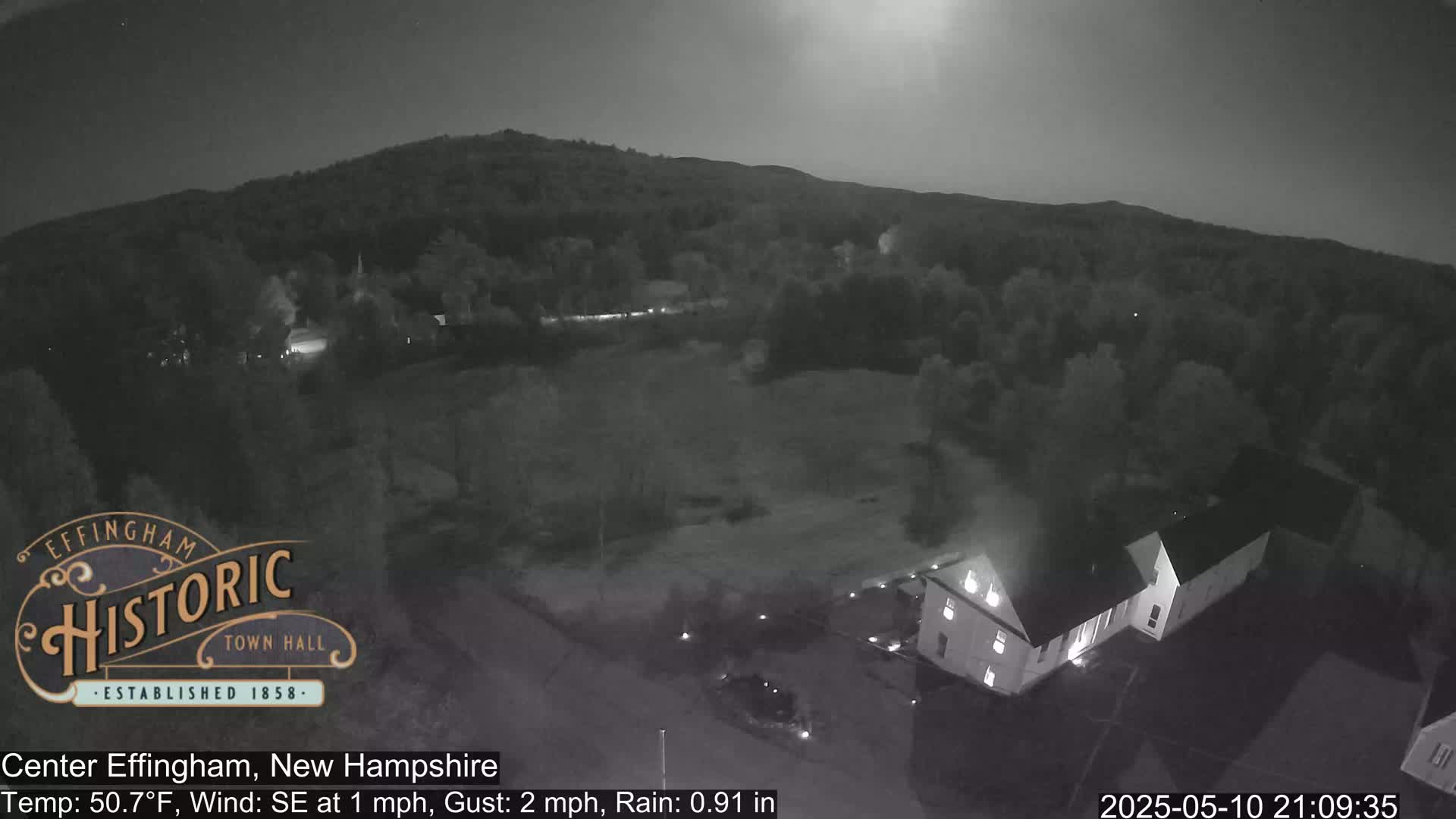 A nighttime, low-light view of a town with a building and trees under a partly cloudy sky, with light rain.