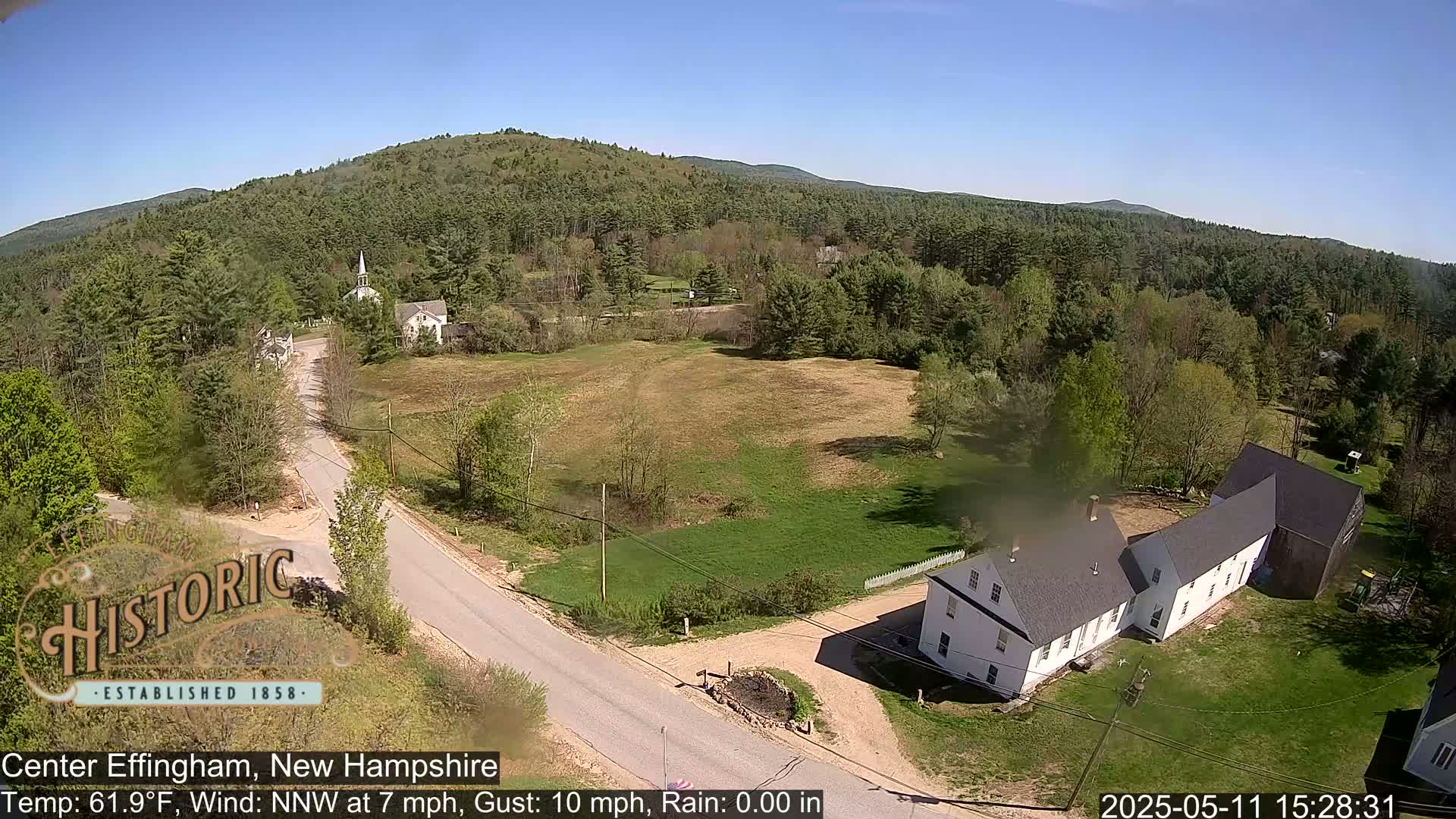An aerial view shows a rural town with a road, buildings, and green fields under a clear blue sky.