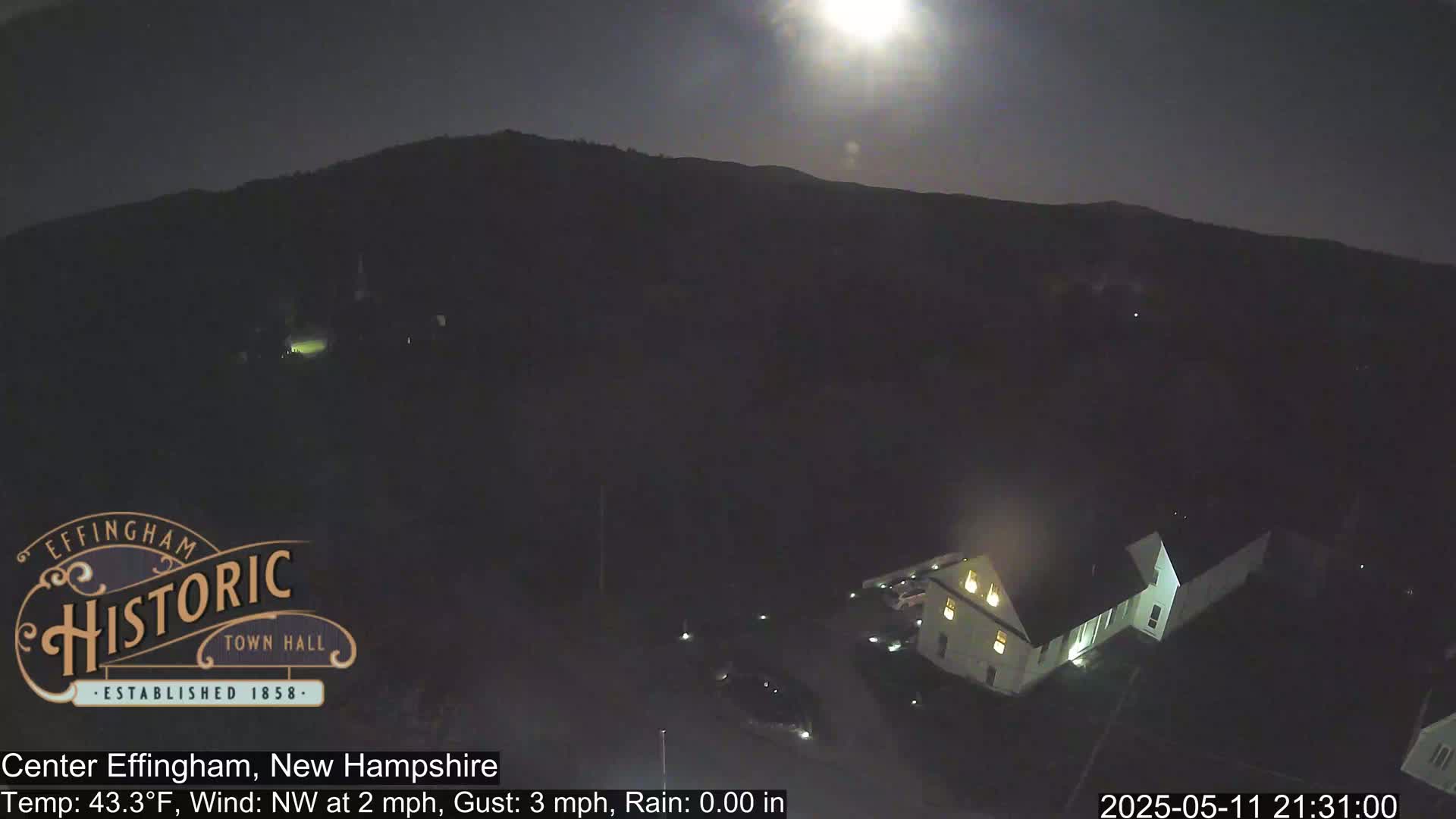 A brightly lit building sits below a dark, moonlit hill under a clear night sky.