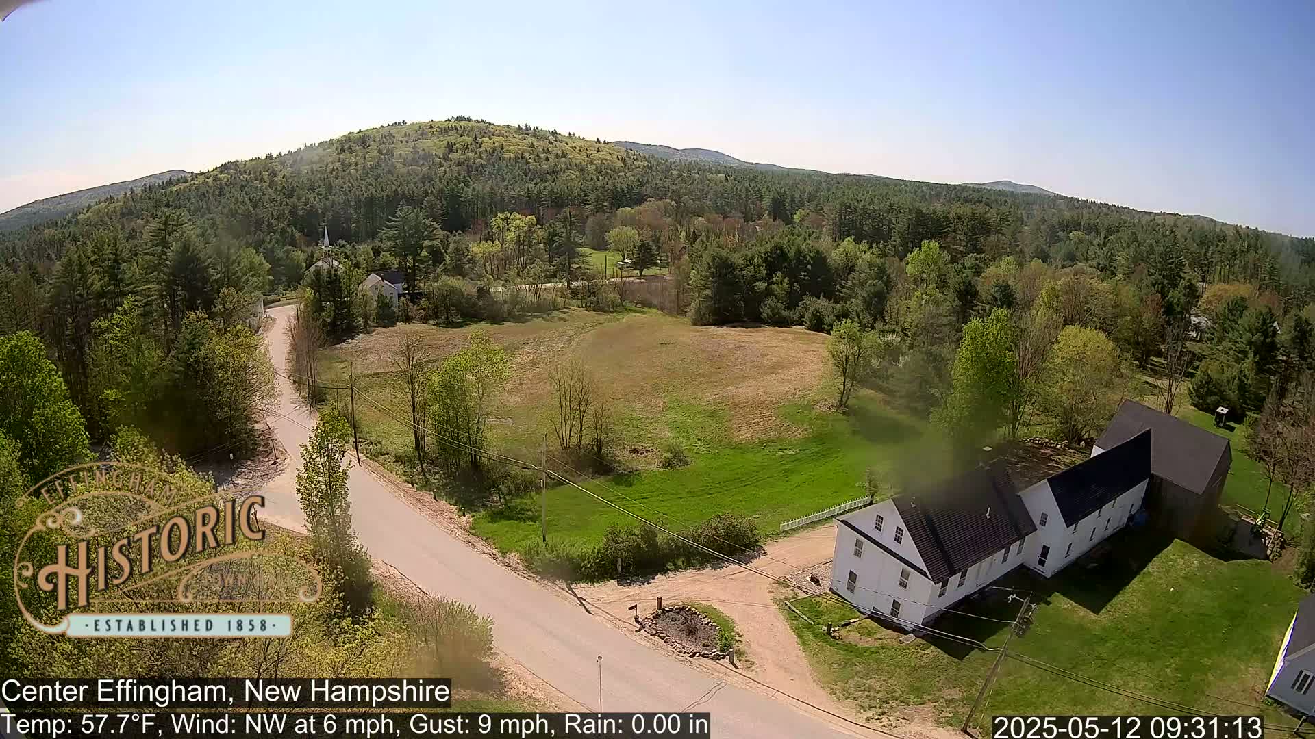 An aerial view shows a rural road intersecting near a white farmhouse and barn, surrounded by green fields and a forested hillside under a clear, sunny sky.