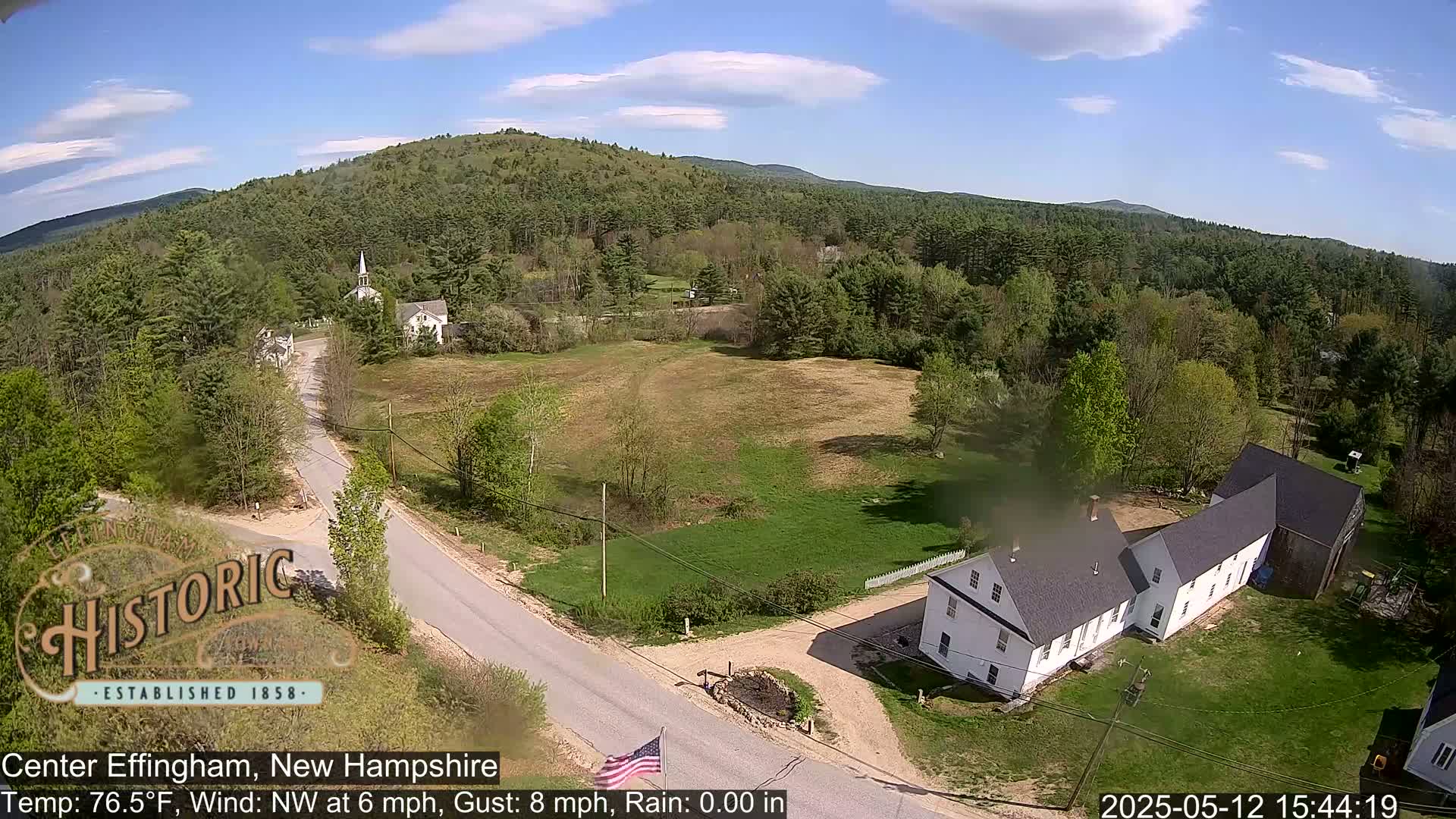 A sunny, slightly windy day reveals a rural New England town with a church steeple, several houses, and green fields, all nestled among lush green trees and hills.