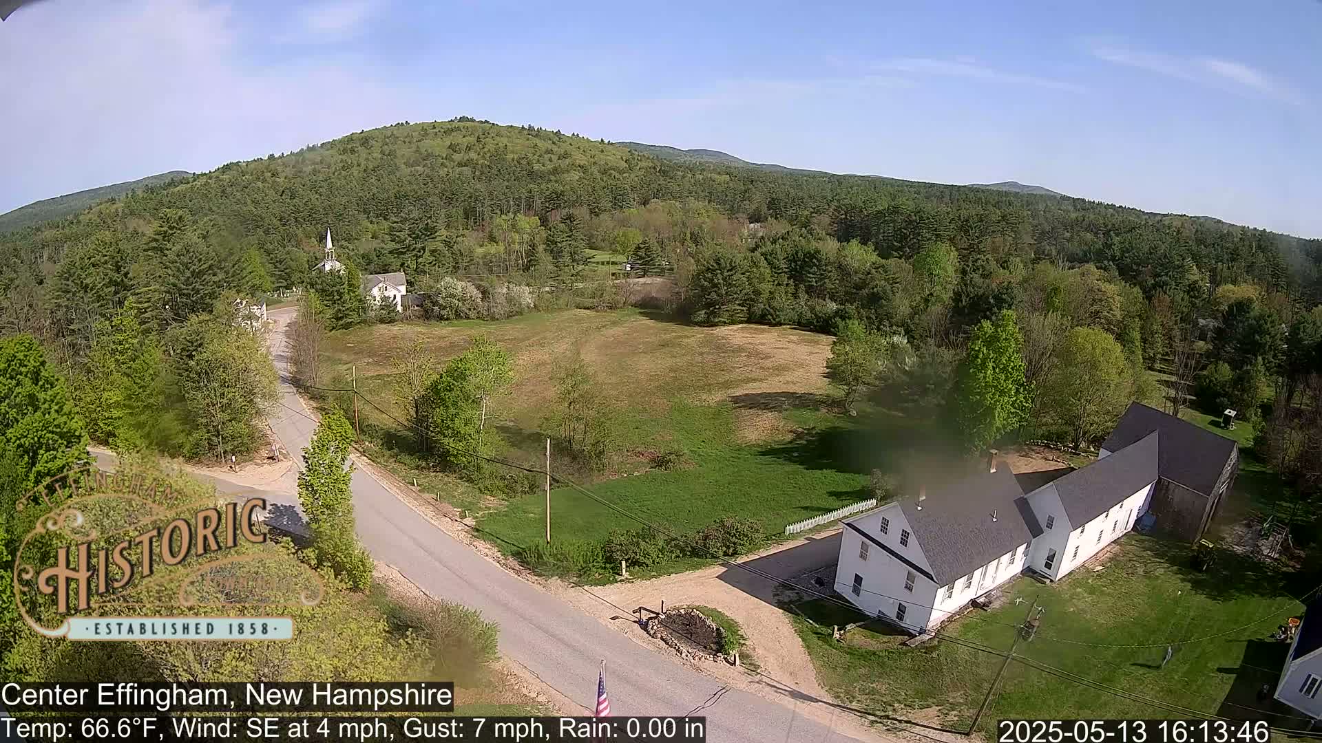 A sunny aerial view of a rural town, showing a road, several buildings, and a large expanse of green fields and trees under a clear blue sky.