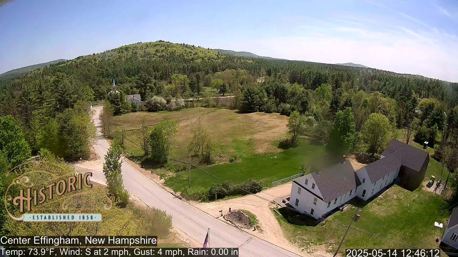 An aerial view of a rural area shows a road intersecting near a white building with a gray roof, surrounded by green trees and fields under a clear, sunny sky.