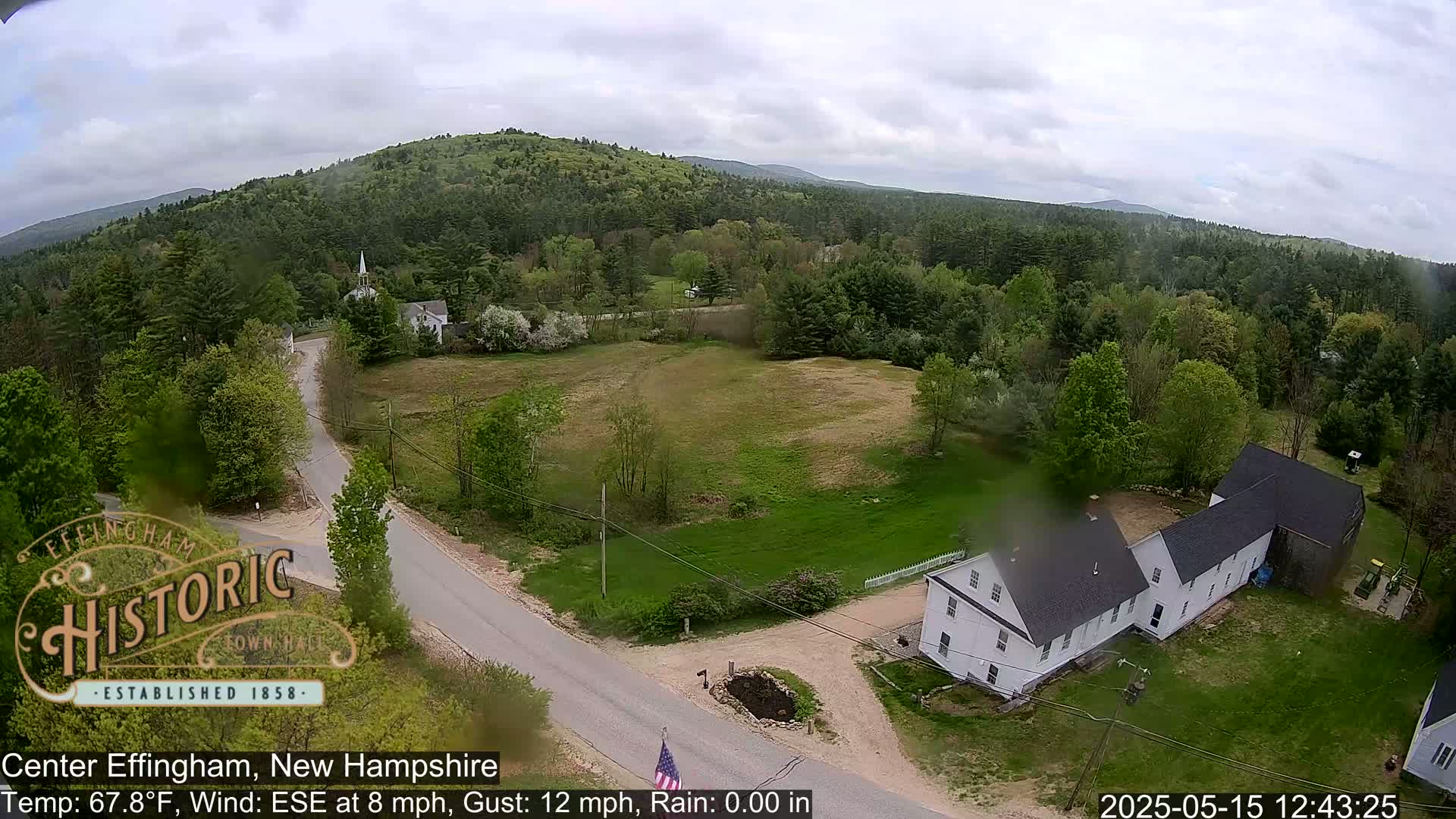 An aerial view shows a rural road intersecting near a white building and a grassy field, surrounded by green trees and hills under a cloudy sky.