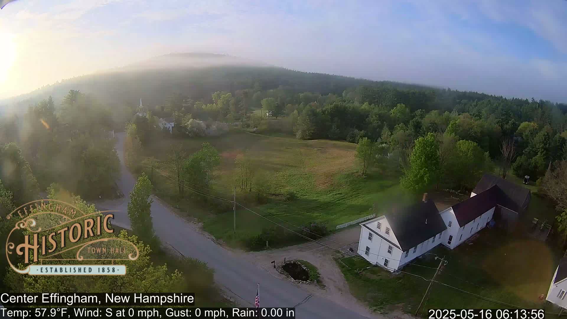 An aerial view shows a rural landscape with a white farmhouse, a road, and green fields under a hazy, calm morning sky.