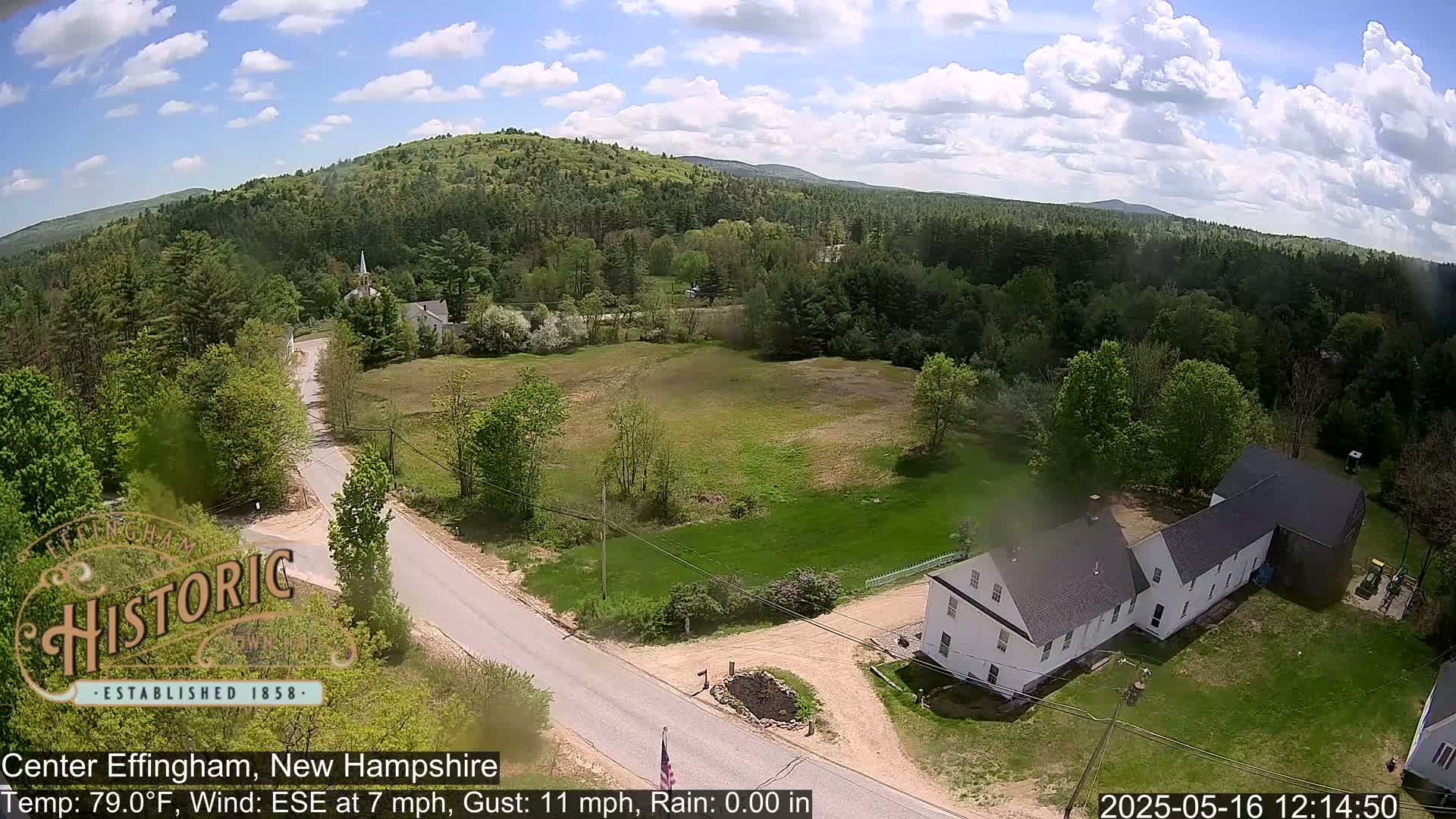 A partly cloudy day reveals a rural New Hampshire landscape featuring a long white building, a road, green fields, and a forested hillside in the background.
