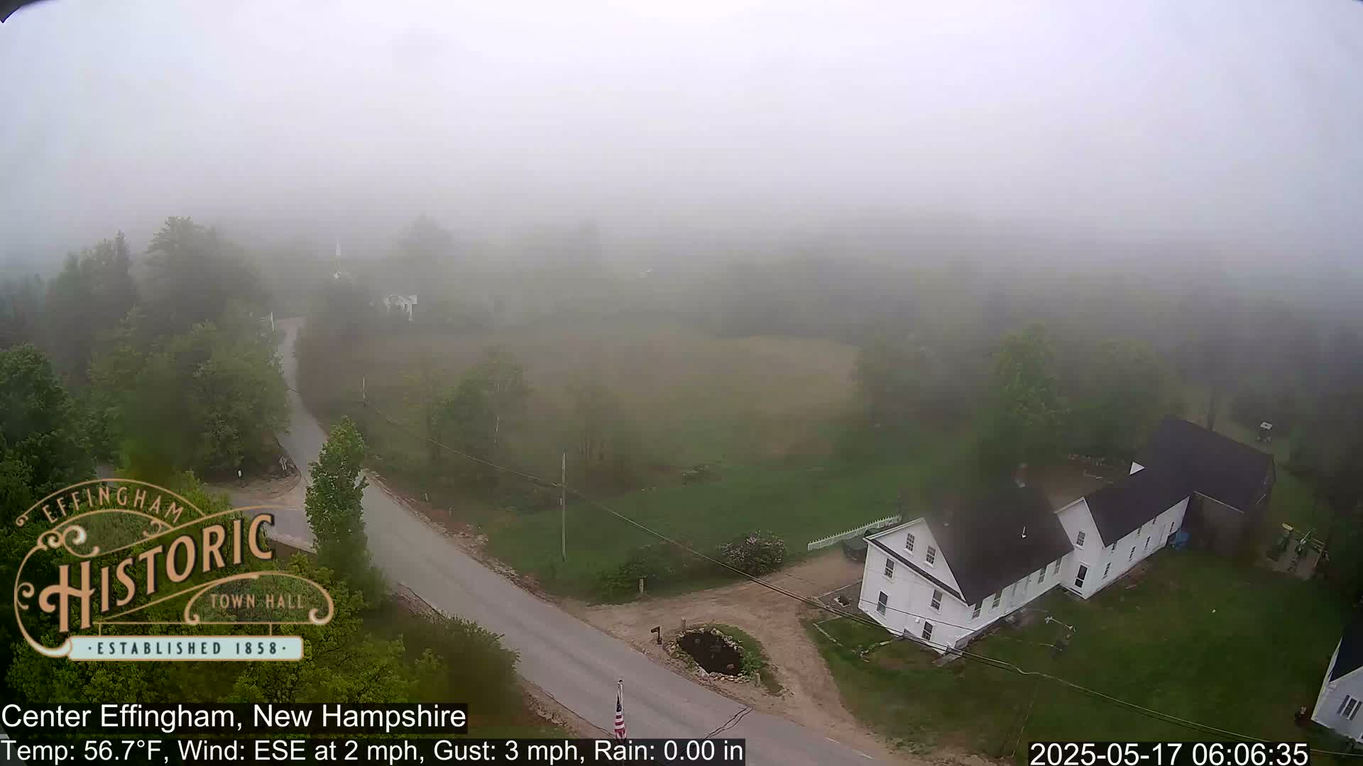 A foggy aerial view shows a rural road intersecting near a white house and other buildings surrounded by green trees and fields.