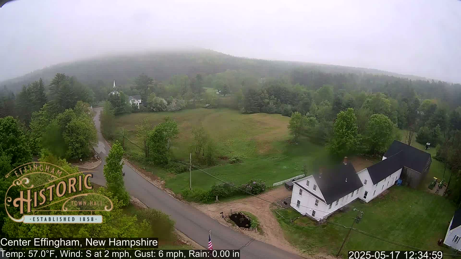 A misty, rainy view of a rural area with a long white house near a road, surrounded by green fields and trees.