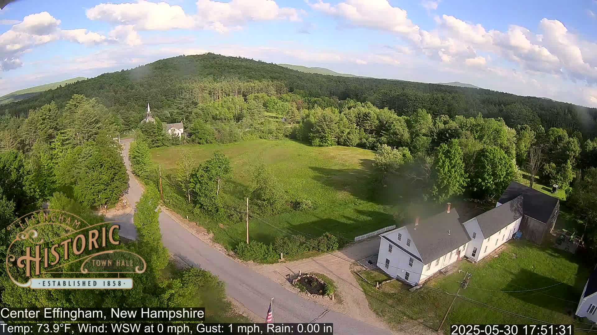 An aerial view shows a partly sunny day over a rural area with green trees, a white building, a church steeple, and roads.