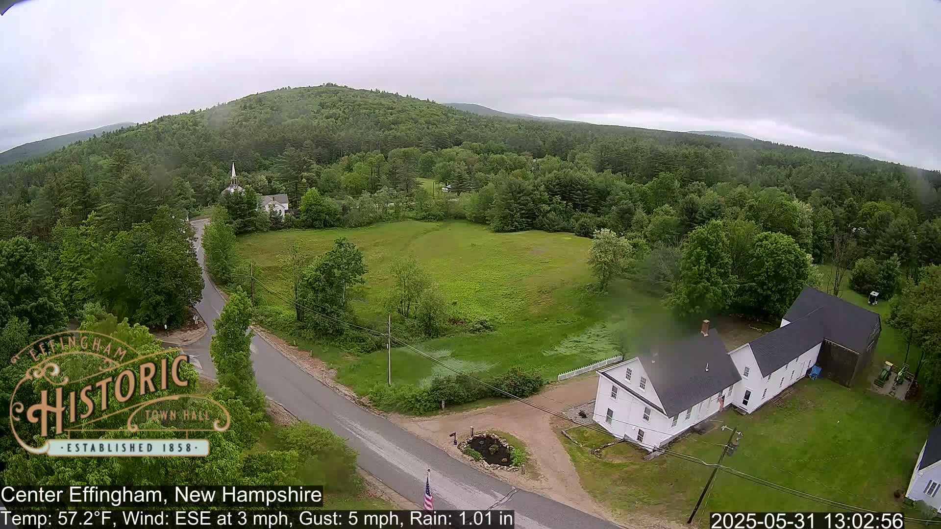A high-angle, rainy view of a rural road intersecting with a grassy field, surrounded by lush green trees and a white house, with a hill in the background.