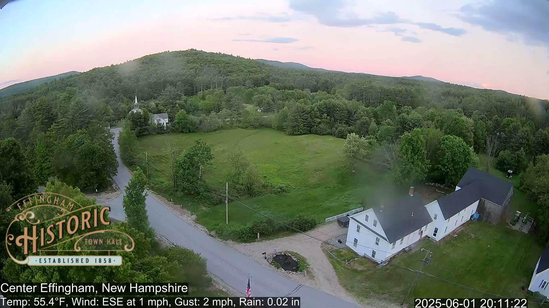A high-angle, long shot of a rural area with a light rain shows a few houses, a church steeple, roads, and green fields, all surrounded by lush green forests and hills under a dusky sky.