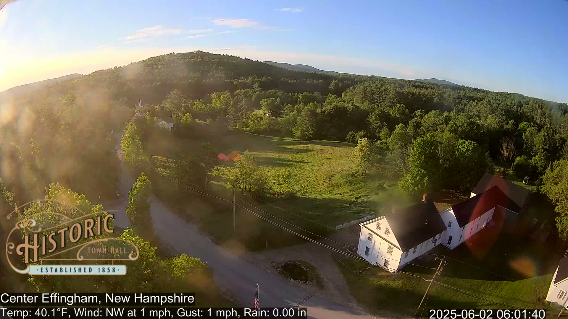 An aerial view shows a rural area with a white house, green fields, and a road, under sunny, clear skies.