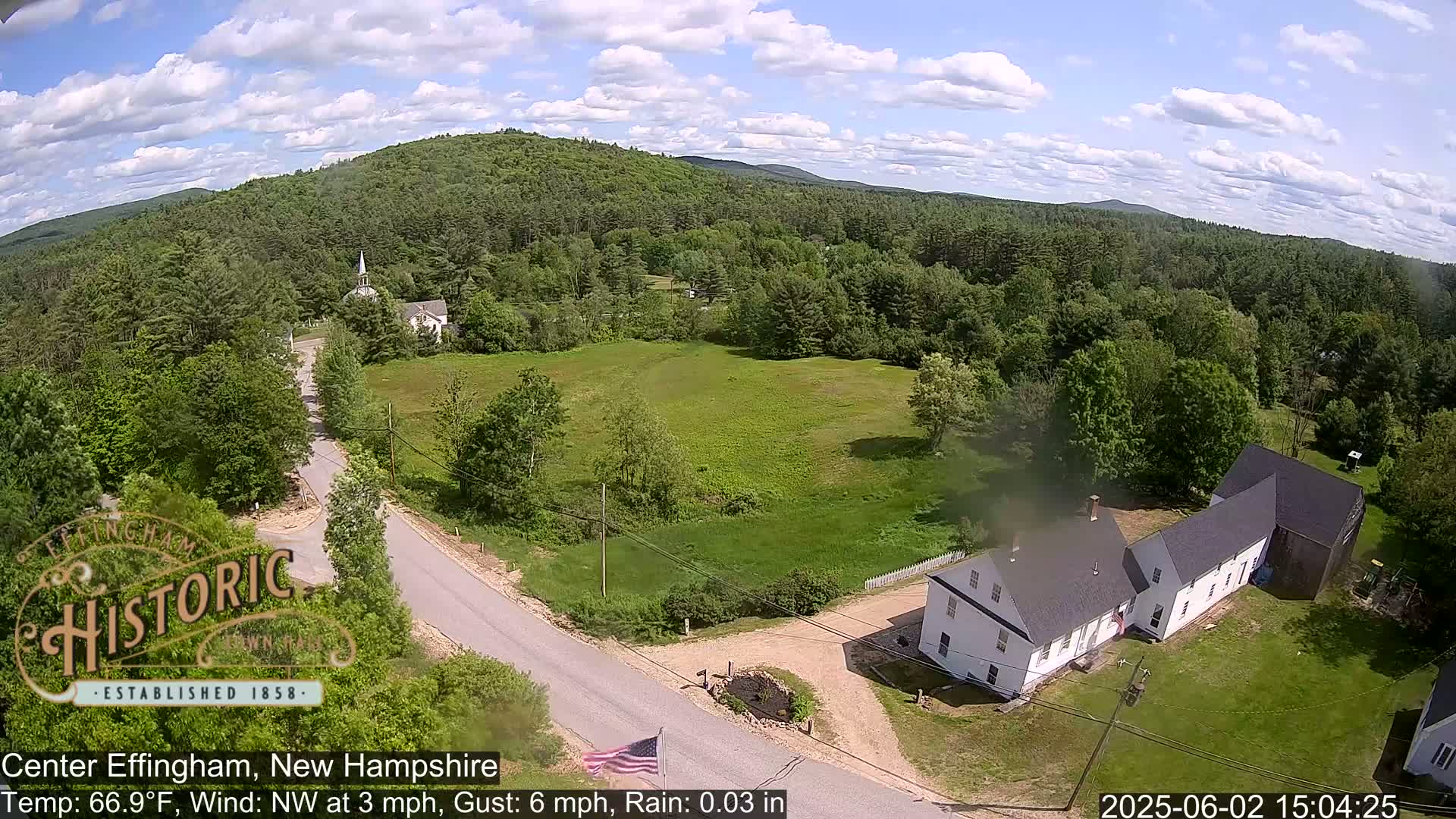 An aerial view shows a rural New England scene under partly cloudy skies, with a road winding through green fields and past houses and a church, nestled among a heavily wooded landscape.