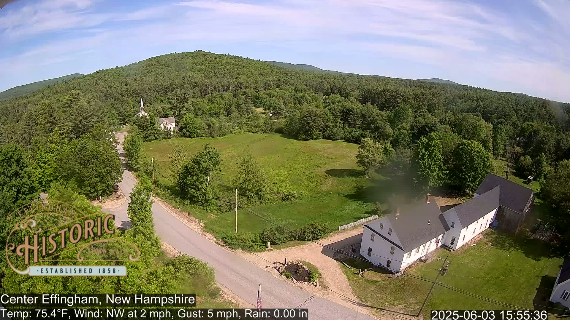 A high-angle view shows a rural intersection with a white building and a church steeple in the distance on a partly sunny day.