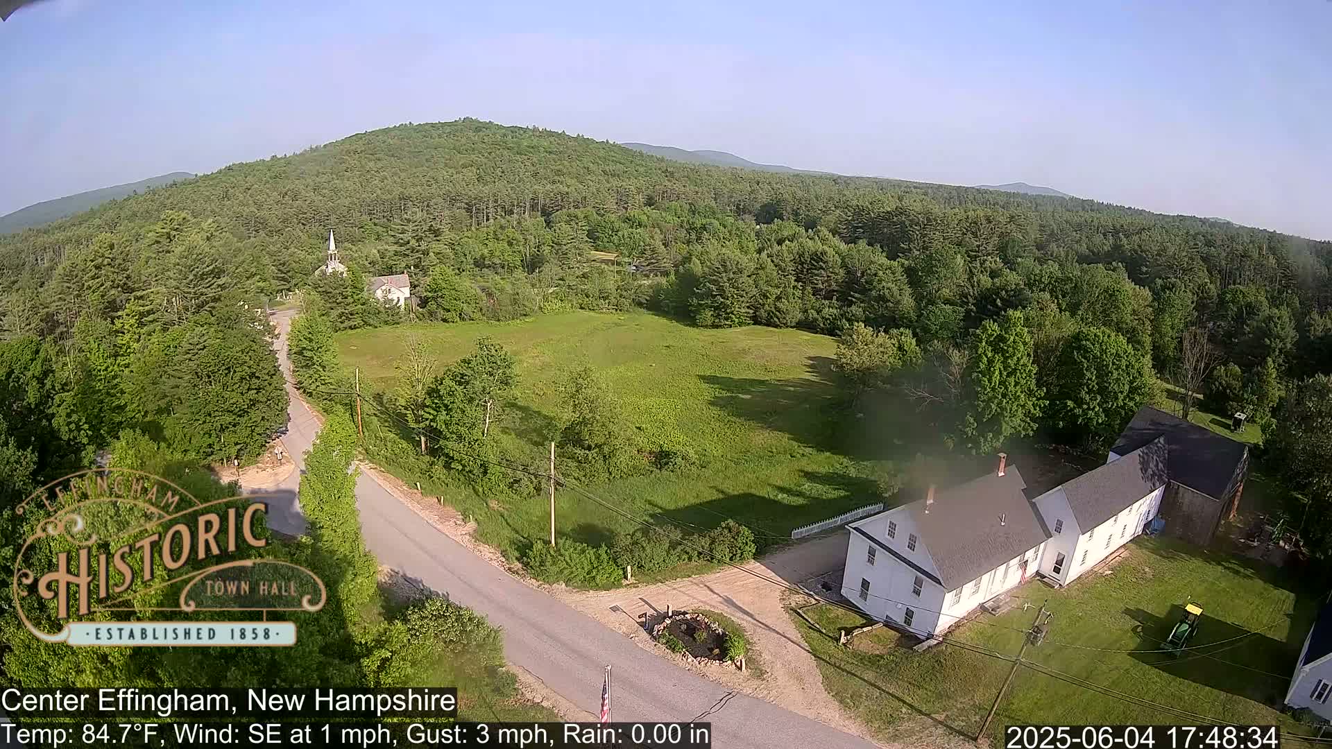 An aerial view shows a small town nestled among green hills and trees on a sunny day with a light southeast wind.