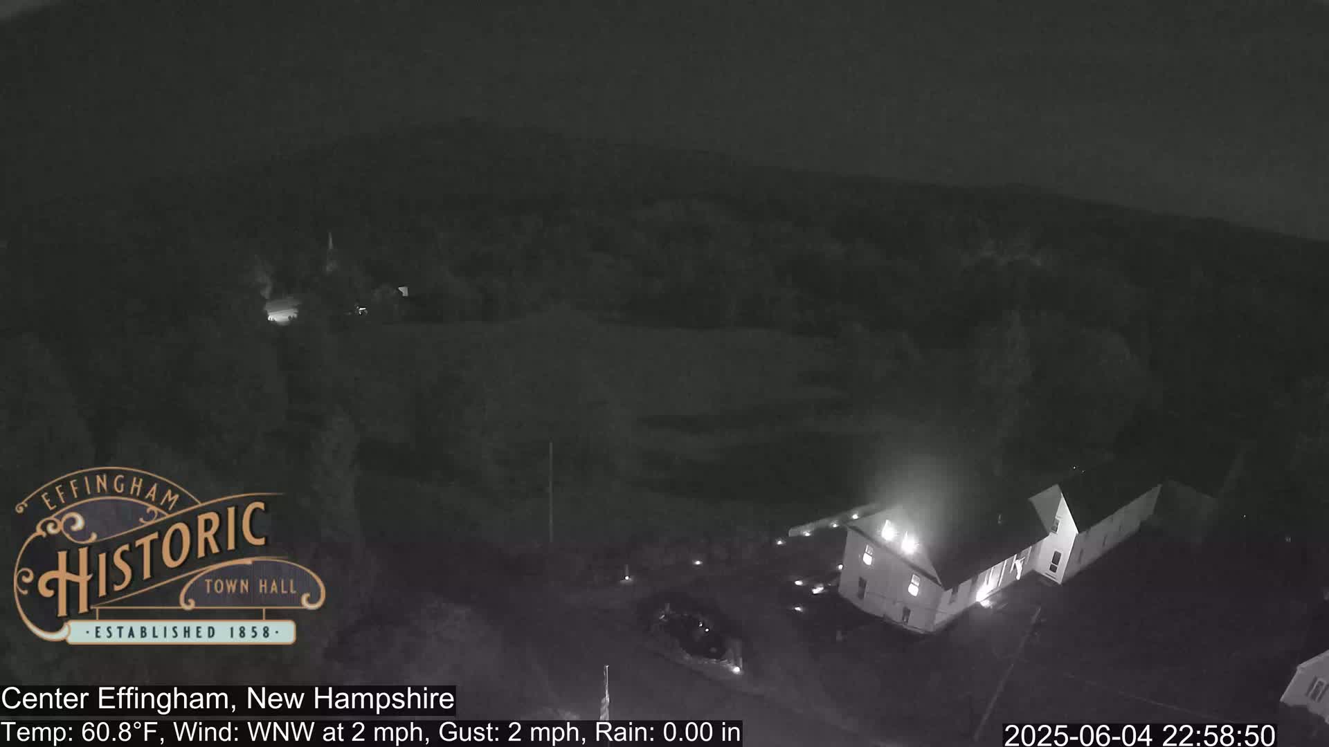 A nighttime, overhead view of a building with lights on, in a dark, rural area with a slight wind.