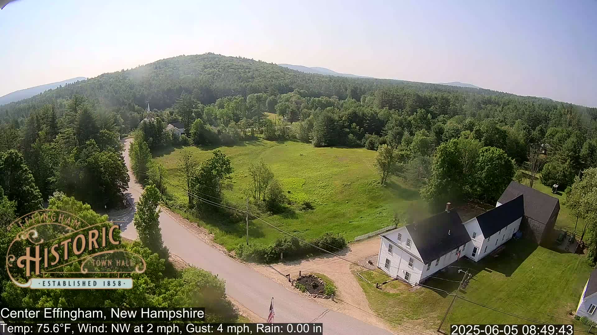 An aerial view shows a rural area with a white farmhouse, a road, green fields, and a forest-covered hill in the background on a clear, sunny day.