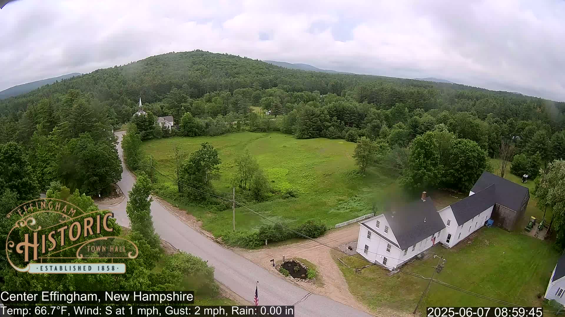 An aerial view shows a rural New Hampshire town on an overcast day, with a white house, a road, and green fields surrounded by a forest and hills.
