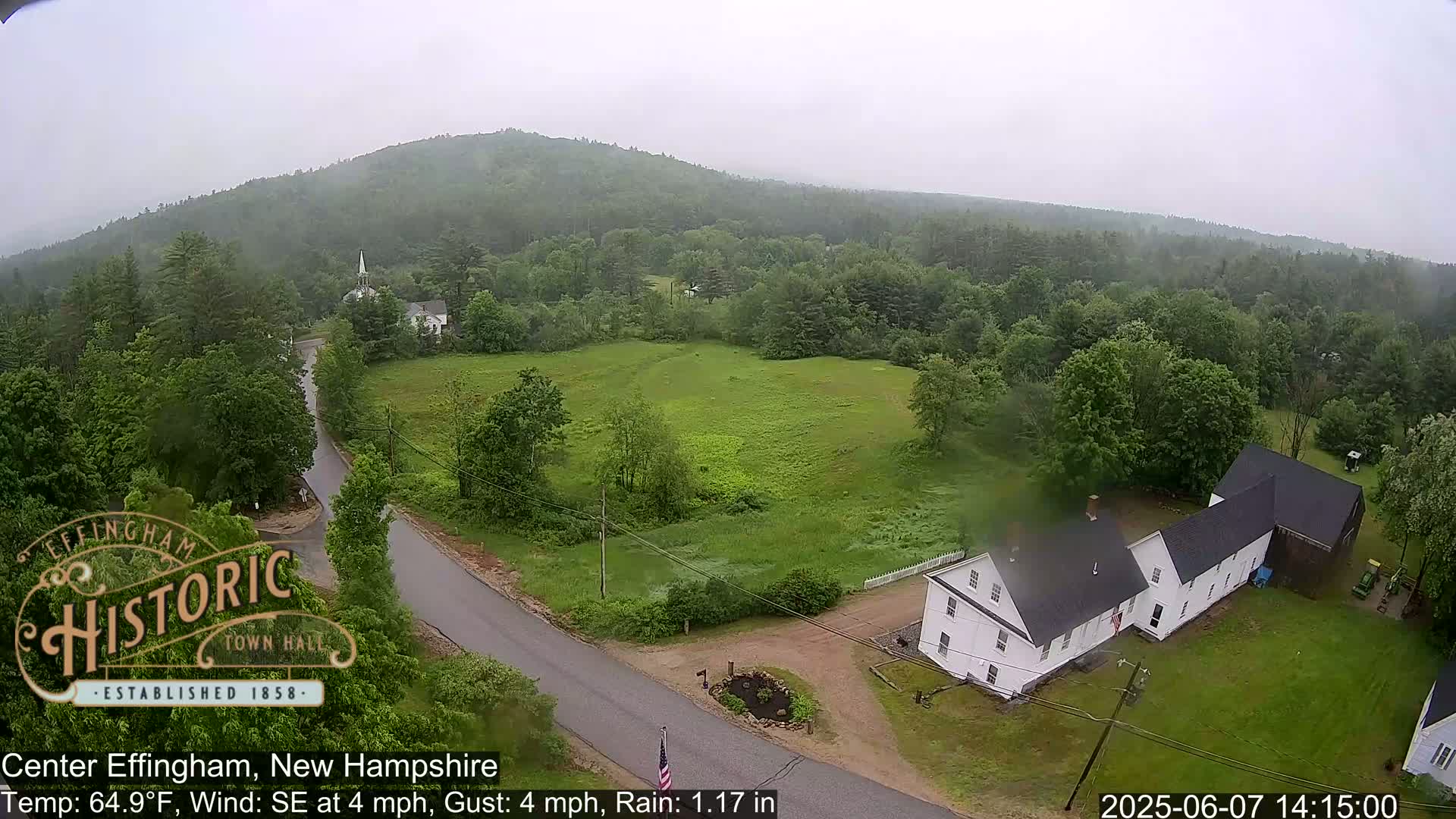 An aerial view shows a rain-soaked rural area with a white house, a road, and lush green fields, all surrounded by a hilly, forested landscape under an overcast sky.
