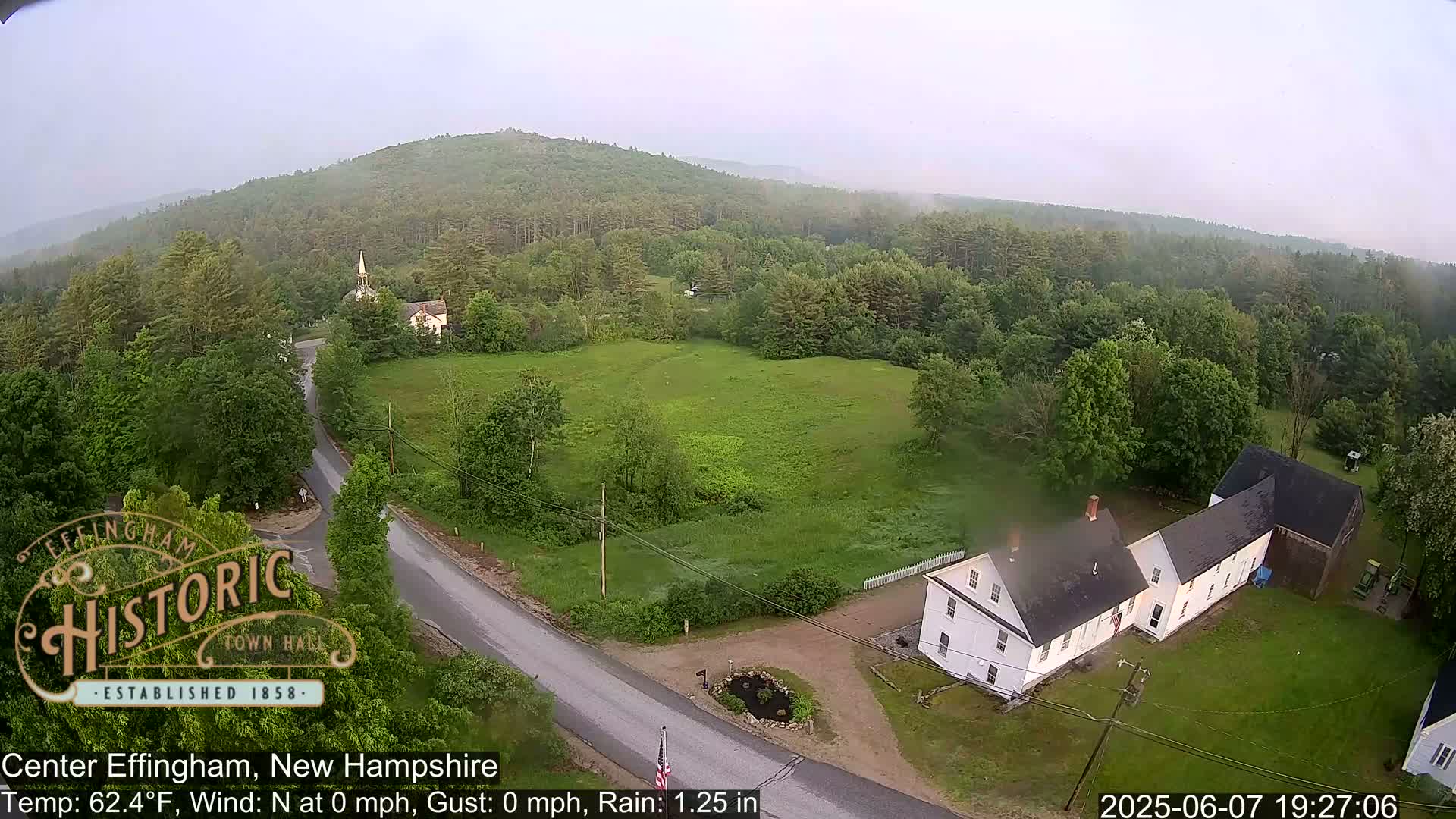 An aerial view shows a rural town in light rain, featuring a road, several buildings, and a hill covered in green trees.
