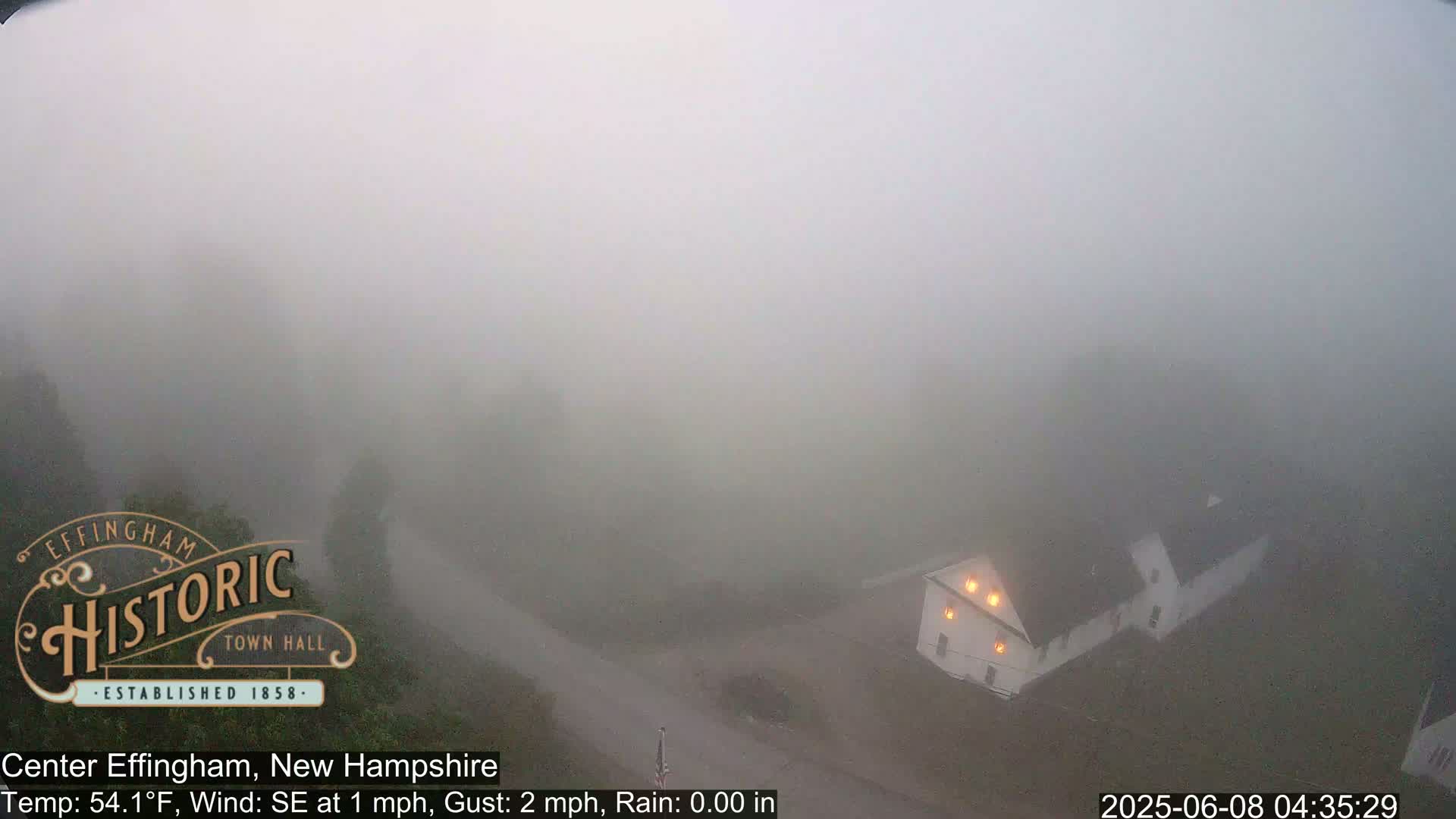 A foggy scene shows a light-colored building with illuminated windows near a road, under a temperature of 54.1°F with a southeast wind of 1 mph and gusts up to 2 mph.
