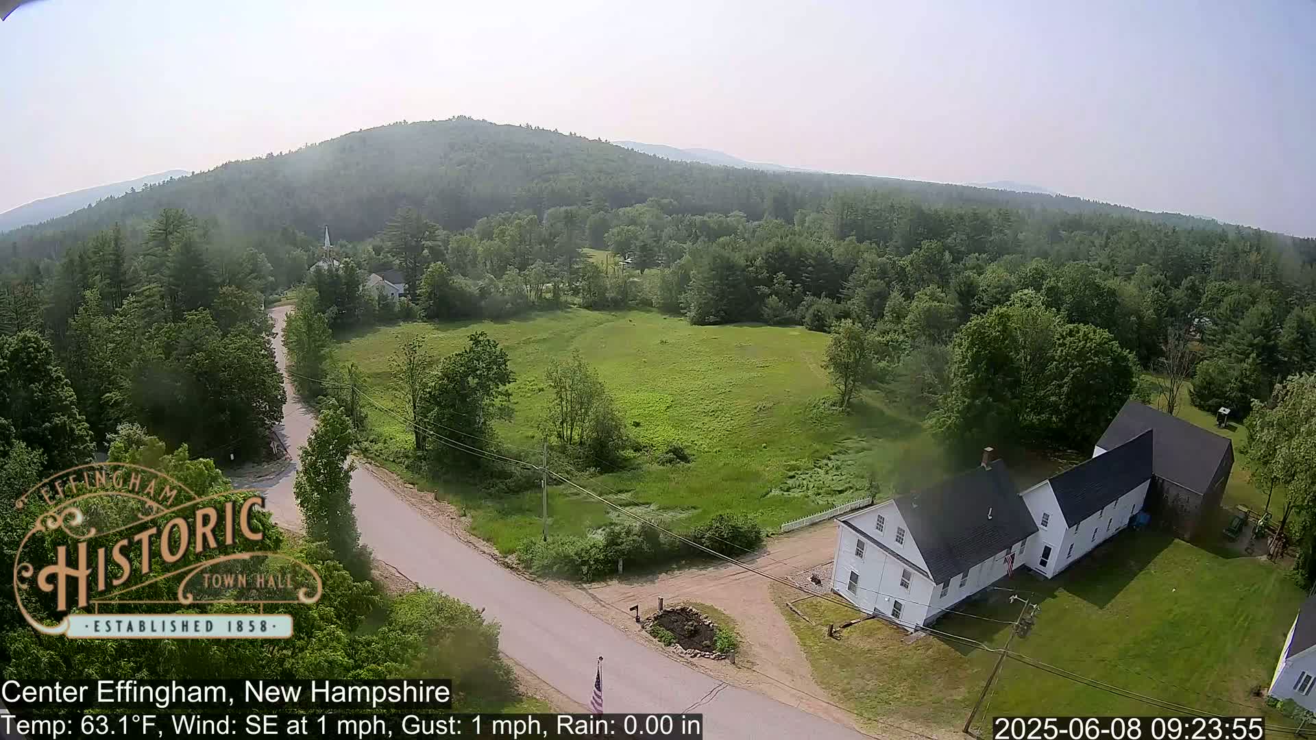 A high-angle, long shot shows a rural New Hampshire landscape with a white building near a road, surrounded by green fields and forest under a hazy sky.