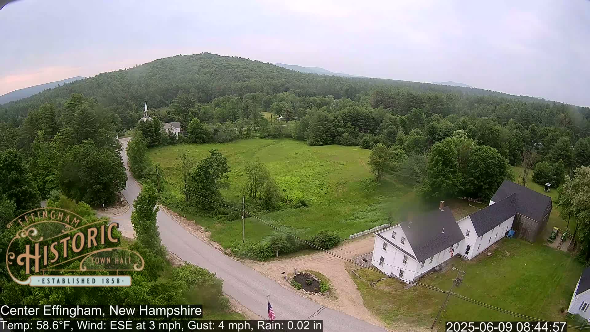 An aerial view shows a rural area with light rain, green fields, a white house, and a road leading towards a church steeple nestled amongst forested hills.