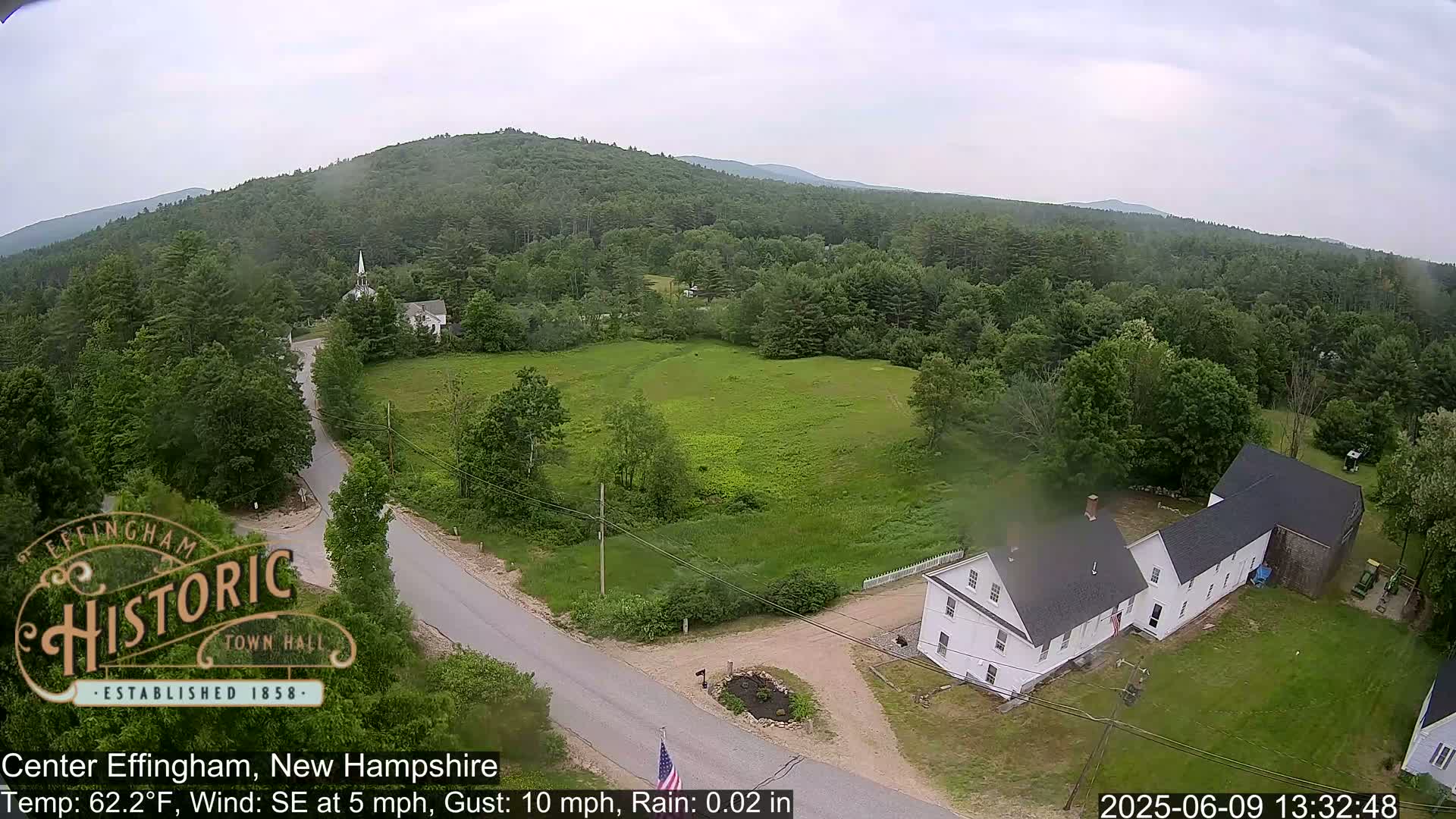 An elevated view of a rural town, showing a road intersection near white buildings and a green field, under overcast skies with light rain.
