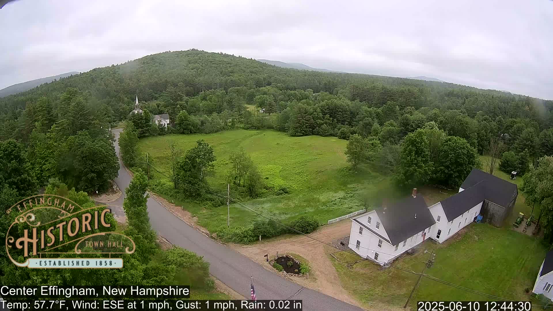 An aerial view shows a lightly rainy day over a rural town nestled amongst green, tree-covered hills, with a white house and a church steeple visible.