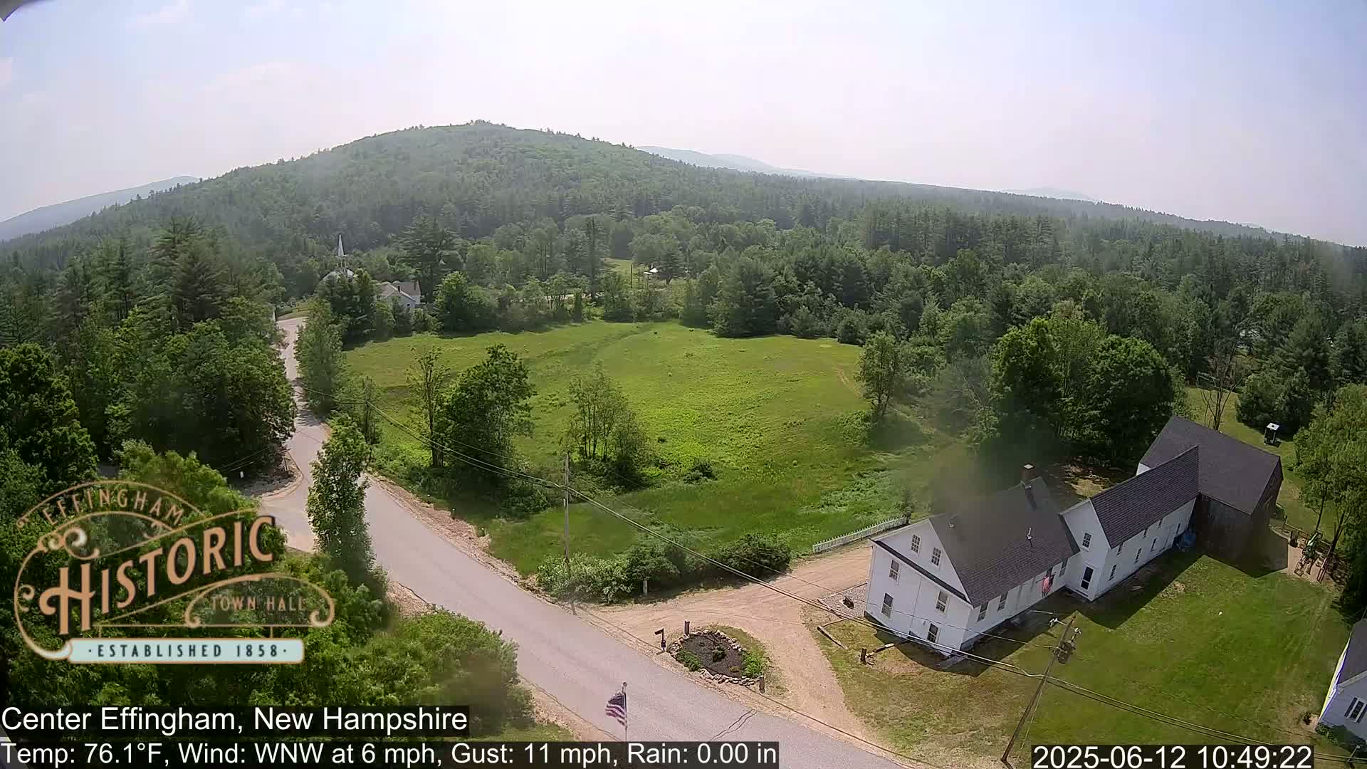 An aerial view shows a rural area with a white farmhouse, a road, green fields, and a forested hill in the background on a partly sunny day with a light wind.