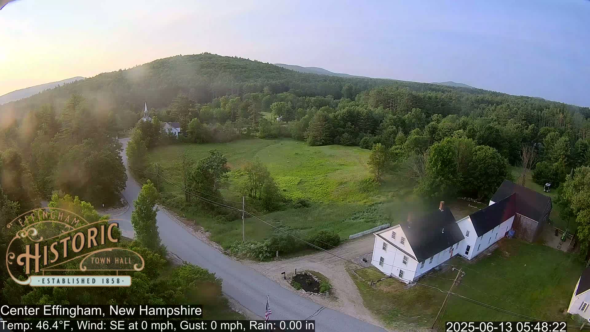 An aerial view shows a rural landscape with a white house, a road, and lush green fields under a clear, light sky.
