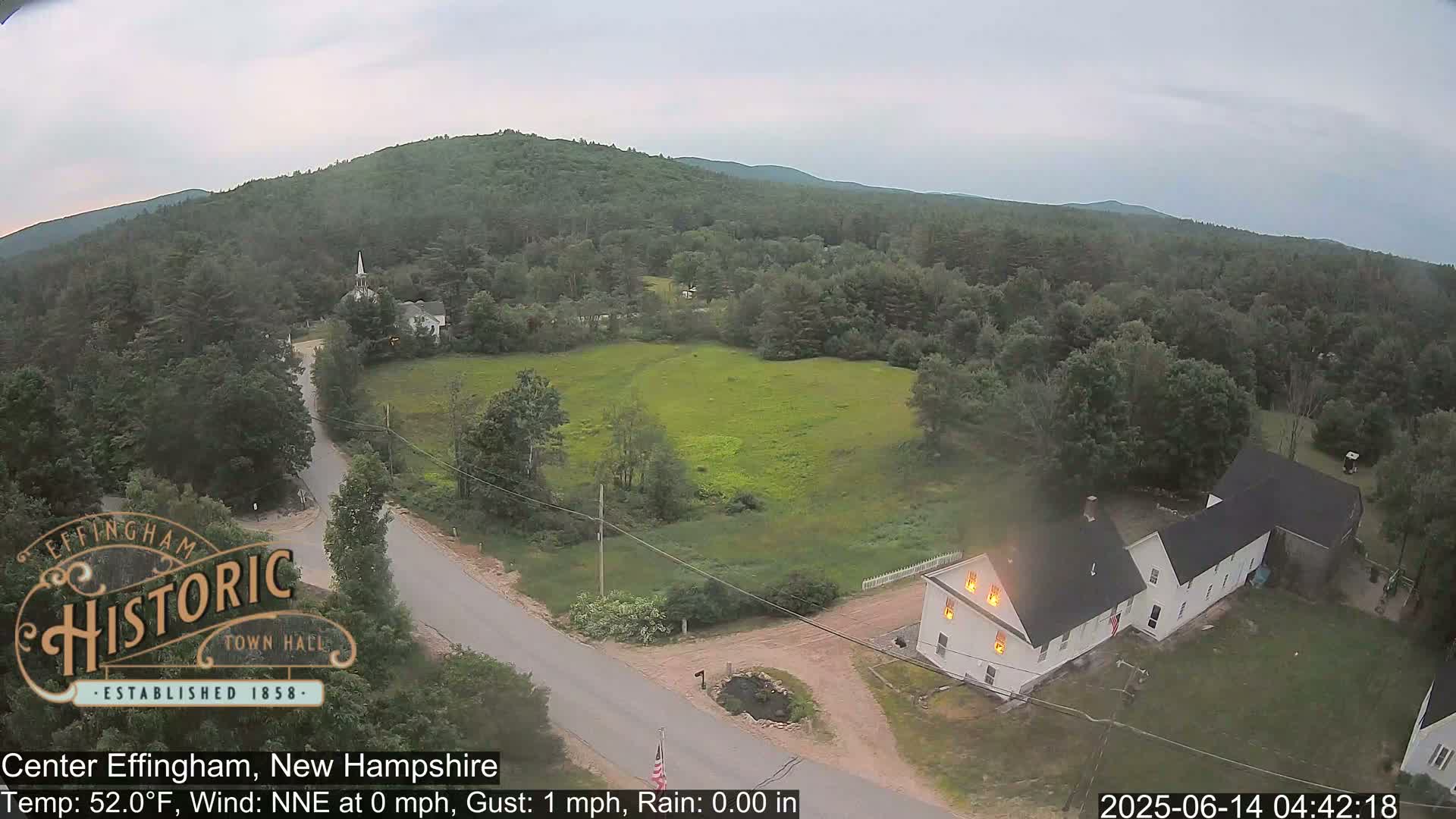 A high-angle view of a rural area shows a house with flames visible in its windows, a road, and green fields under an overcast sky.