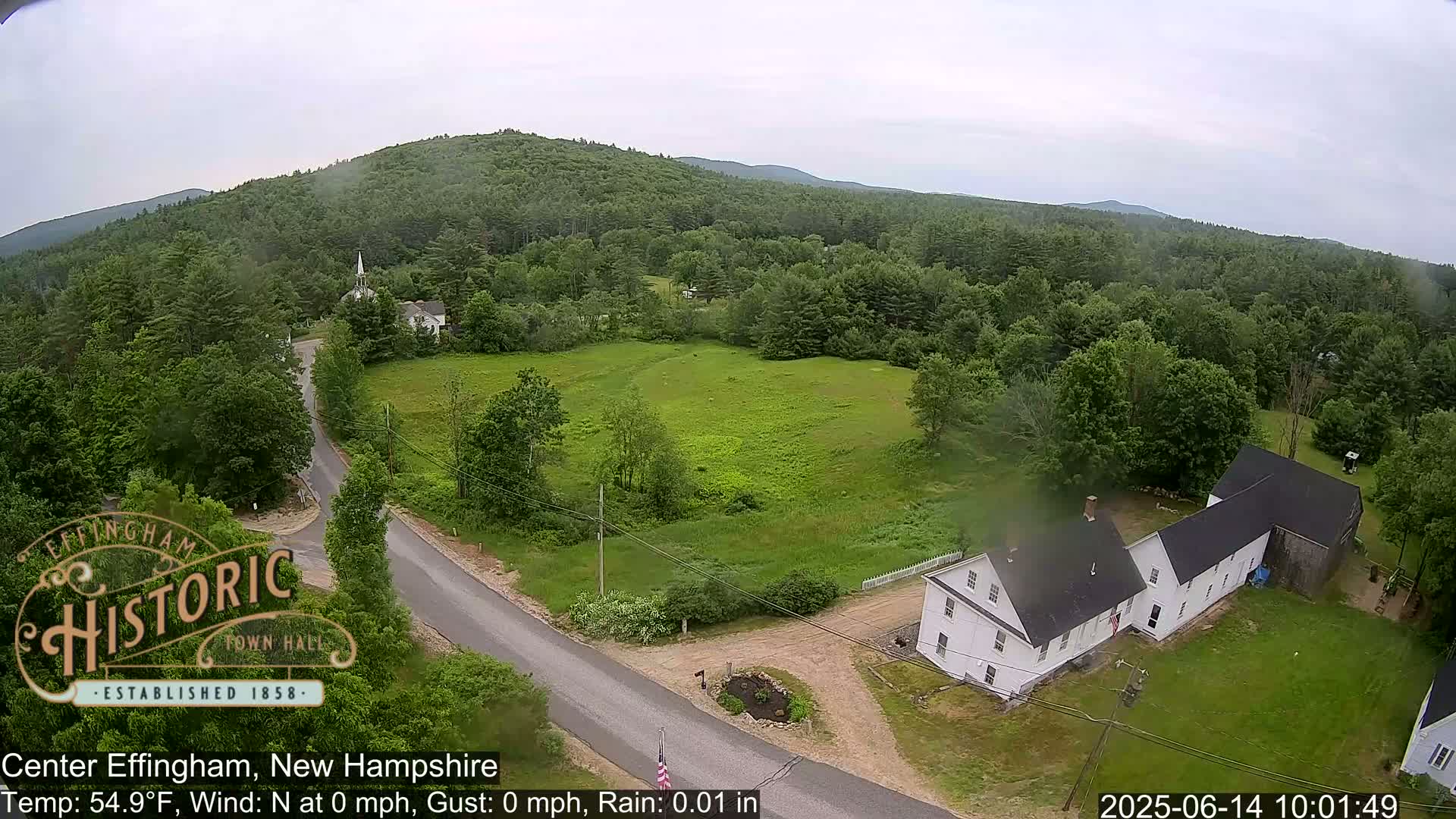An aerial view shows a light rain falling over a rural town nestled among green, forested hills.