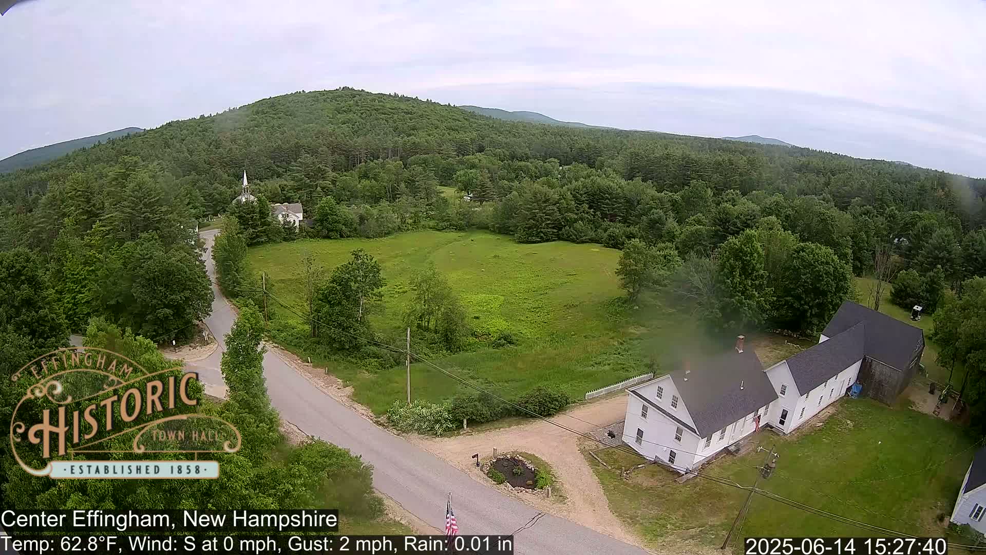 An aerial view shows a light rain falling over a rural town nestled among green, forested hills.