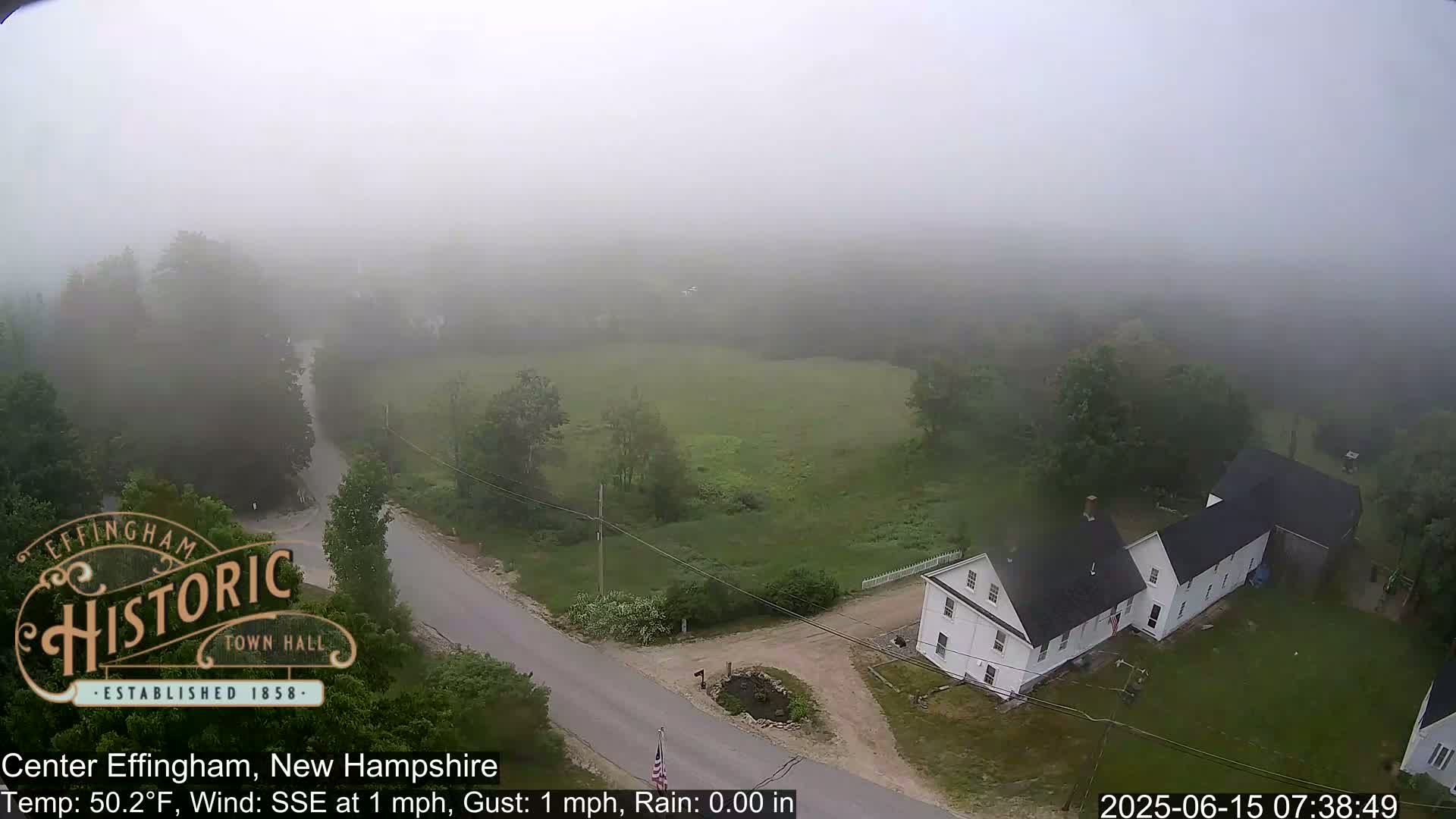 A foggy aerial view shows a rural road intersecting with another road near a white house, with a field and trees visible in the distance.