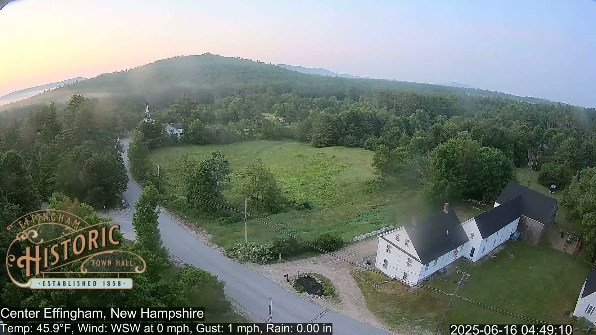 A high-angle view of a rural landscape with low-lying fog, showing a road, a white house, fields, and a forested hillside under a clear, pale sky.