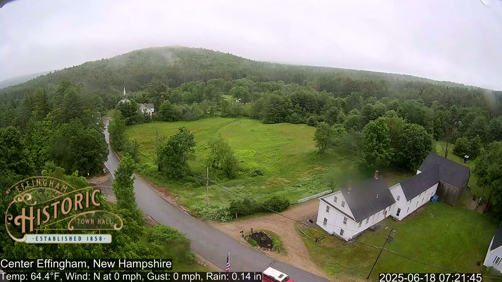 A high-angle view shows a small town nestled in a verdant, forested valley under a lightly overcast sky with a light rain.