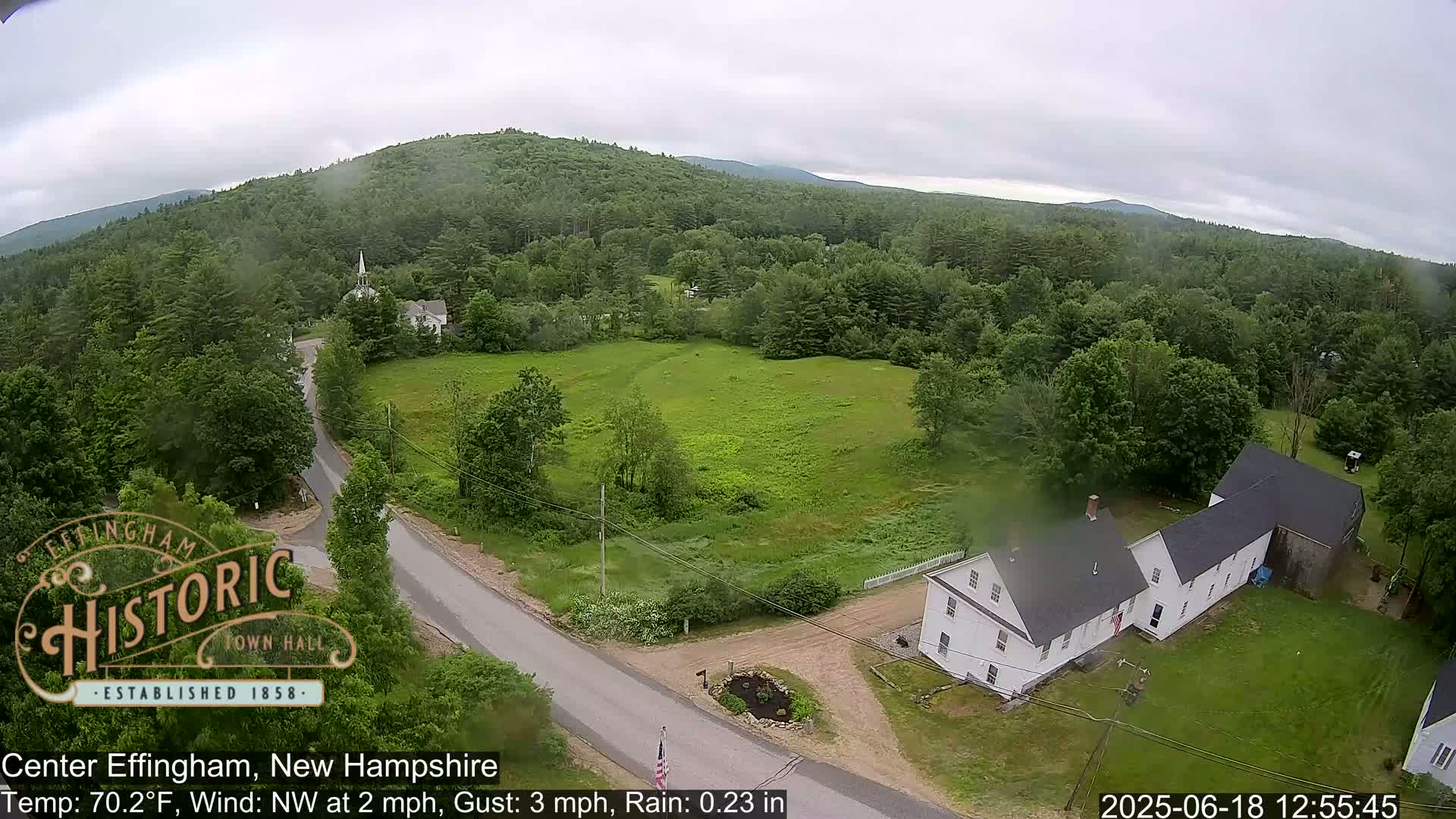 An aerial view of a rural town in rainy weather shows a long white building, roads, a grassy field, and a forest-covered hill in the background.